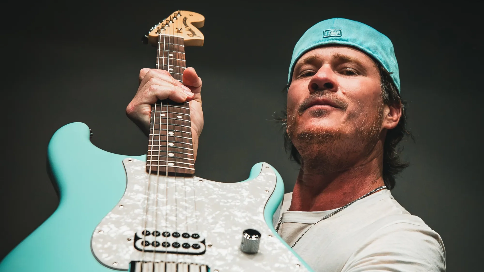 Tom Delonge holding his turquoise Stratocaster signature guitar, wearing a light blue hat and shirt.