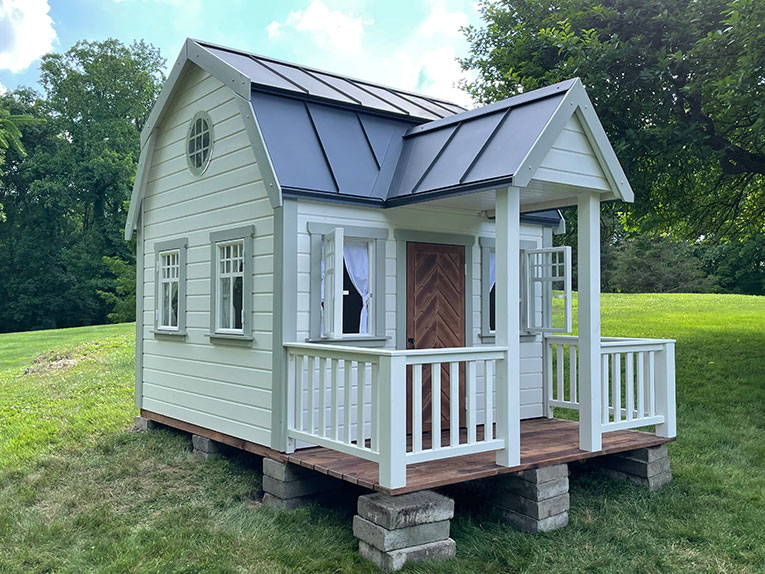 Outside of Kids Playhouse Grand Farmhouse black and white Outdoor Playhouse With Wooden Porch And White Railing, Brown Fishbone Wooden Door by WholeWoodPlayhouses