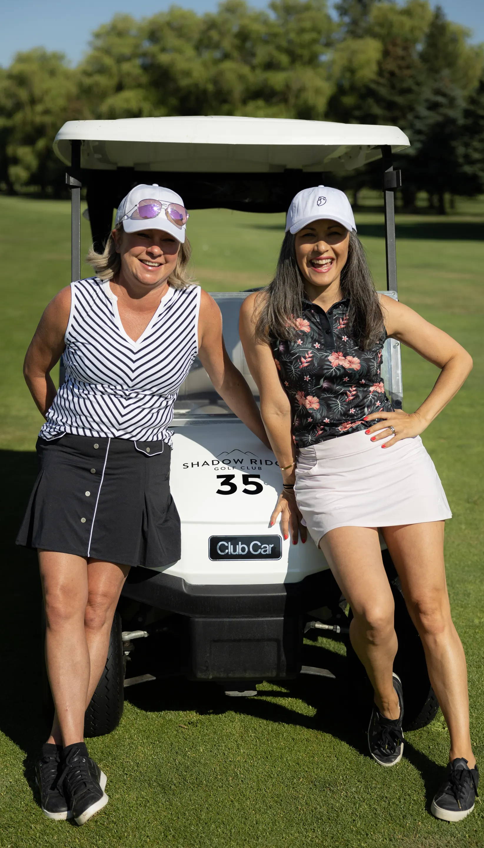 Two female golf entrepreneurs in golf apparel lean against a golf cart on a golf coure in summer.