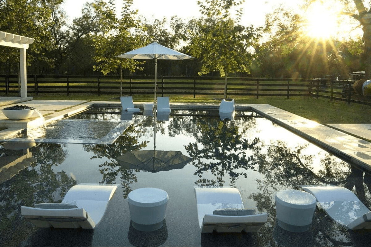 In-pool loungers and round side tables on a shallow tanning ledge, with a center umbrella and sunset light reflecting on the pool surface.