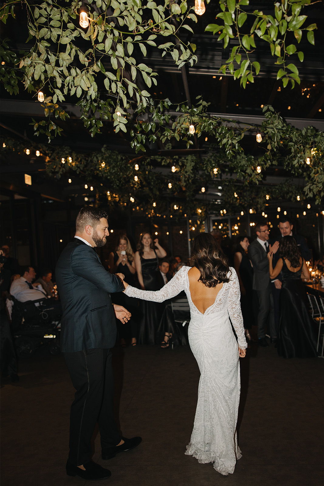 A couple dances at a wedding reception under hanging lights and greenery.