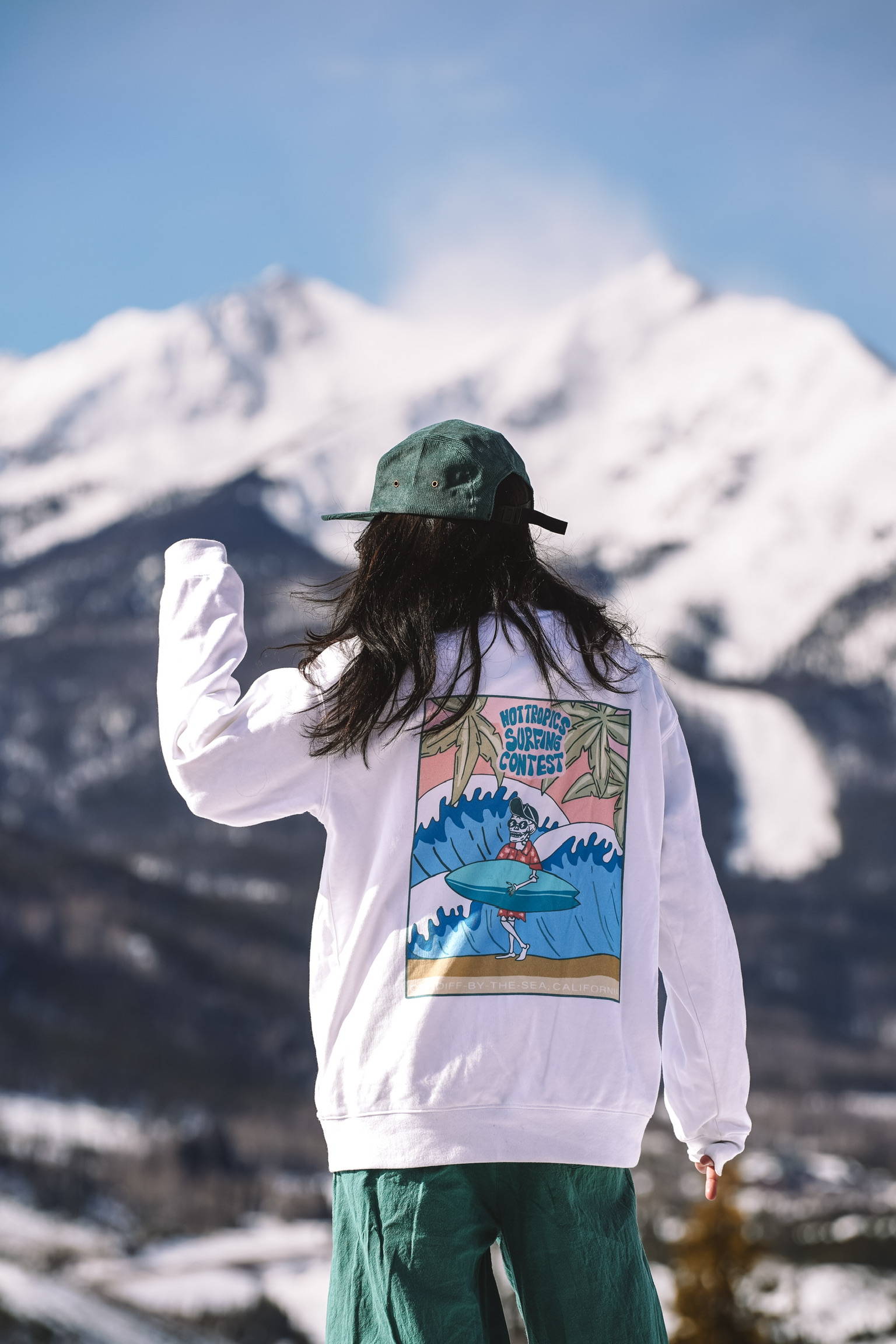 A woman standing in front of mountains wearing a white crewneck and green cap