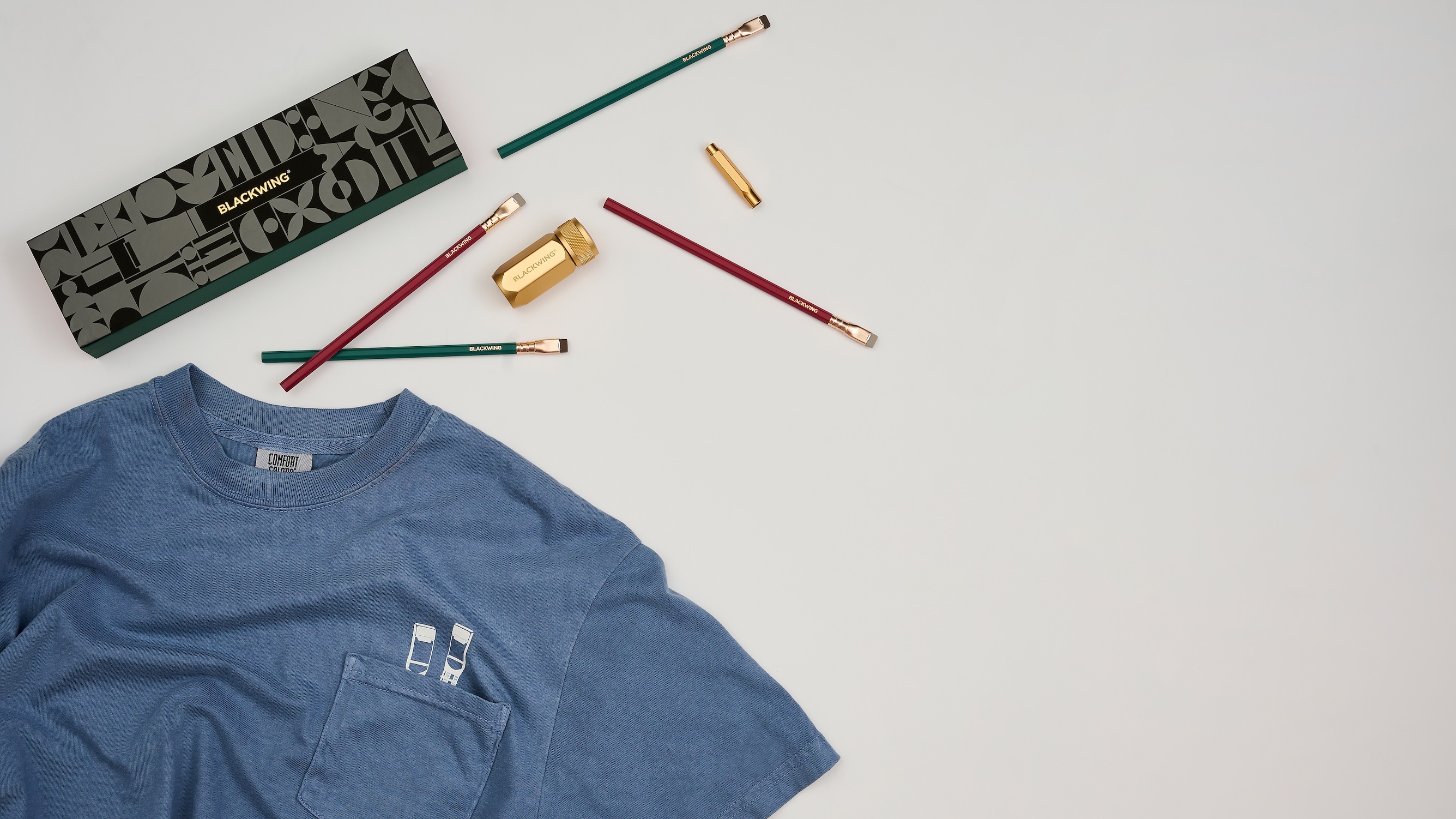 A blue pocket t-shirt with white pencil tops screenprinted over top of pocket, which sits next to 2 green and 2 red pencils, a gold sharpener, a gold point guard, and a black gift box that says 'BLACKWING' all on a white background