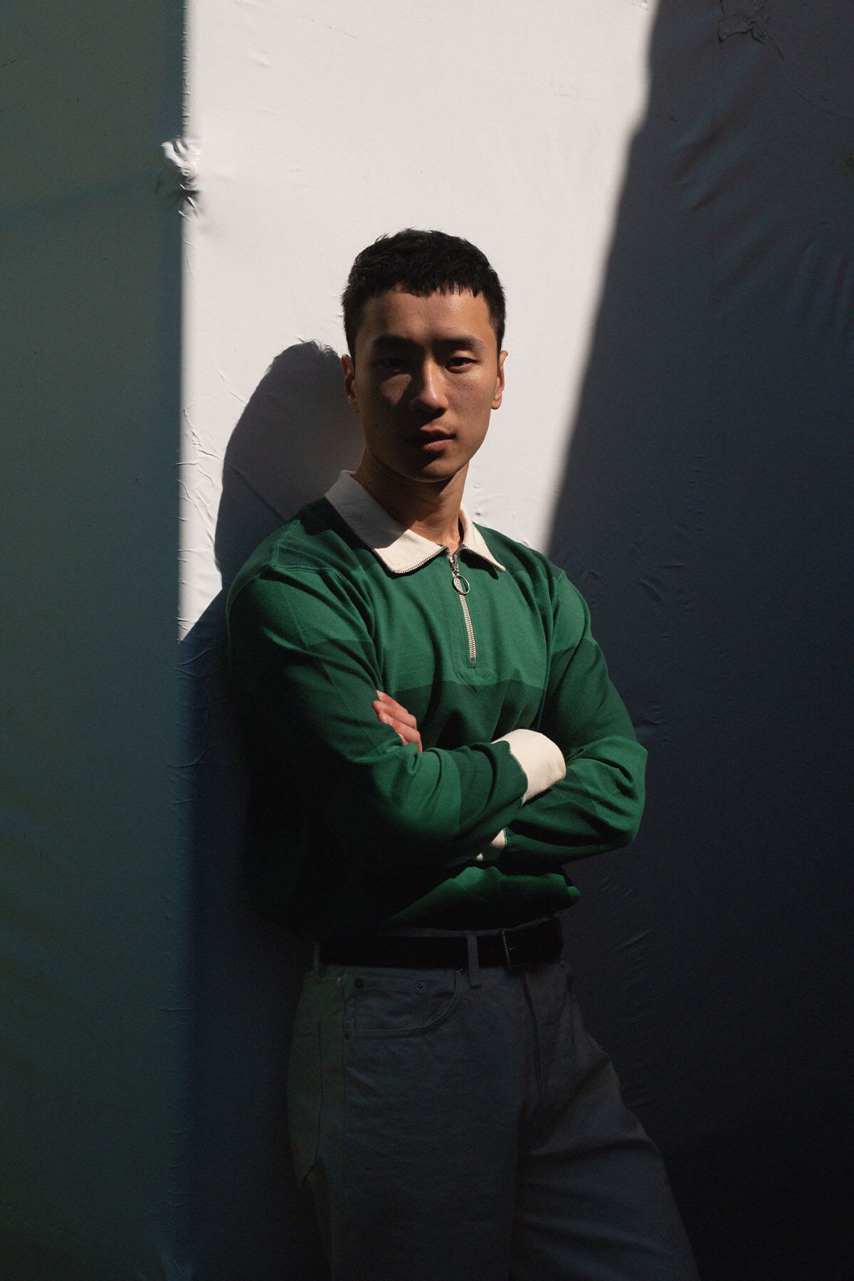 A person in a green sweater stands against a wall, arms crossed.