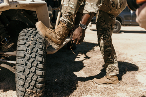 Service member kneeling by a military vehicle tire, lacing a coyote tactical boot