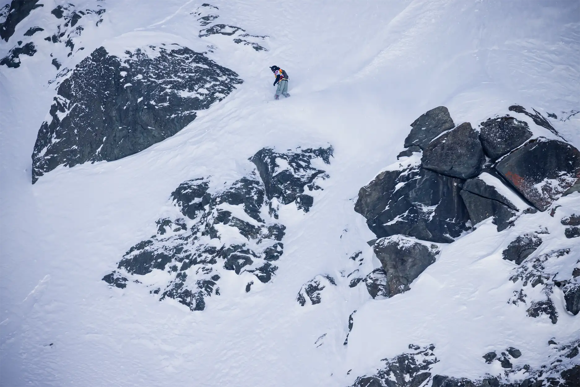 Snowboarder descending snowy mountain with rocky outcrops.