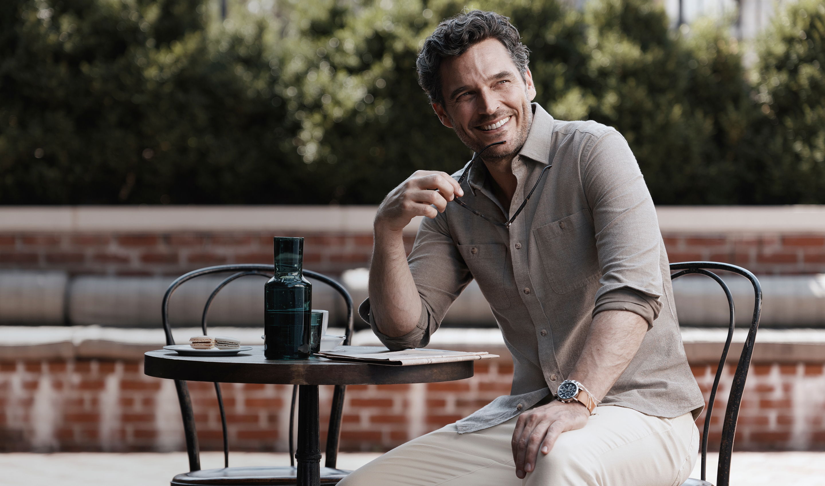 Man sitting at a table outdoors, smiling and holding a drink.