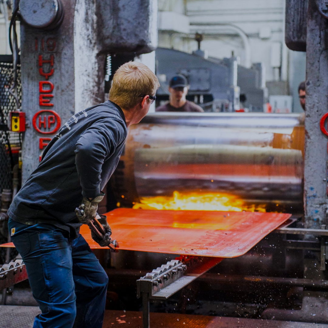 Worker operating machinery with glowing metal sheet in industrial setting.