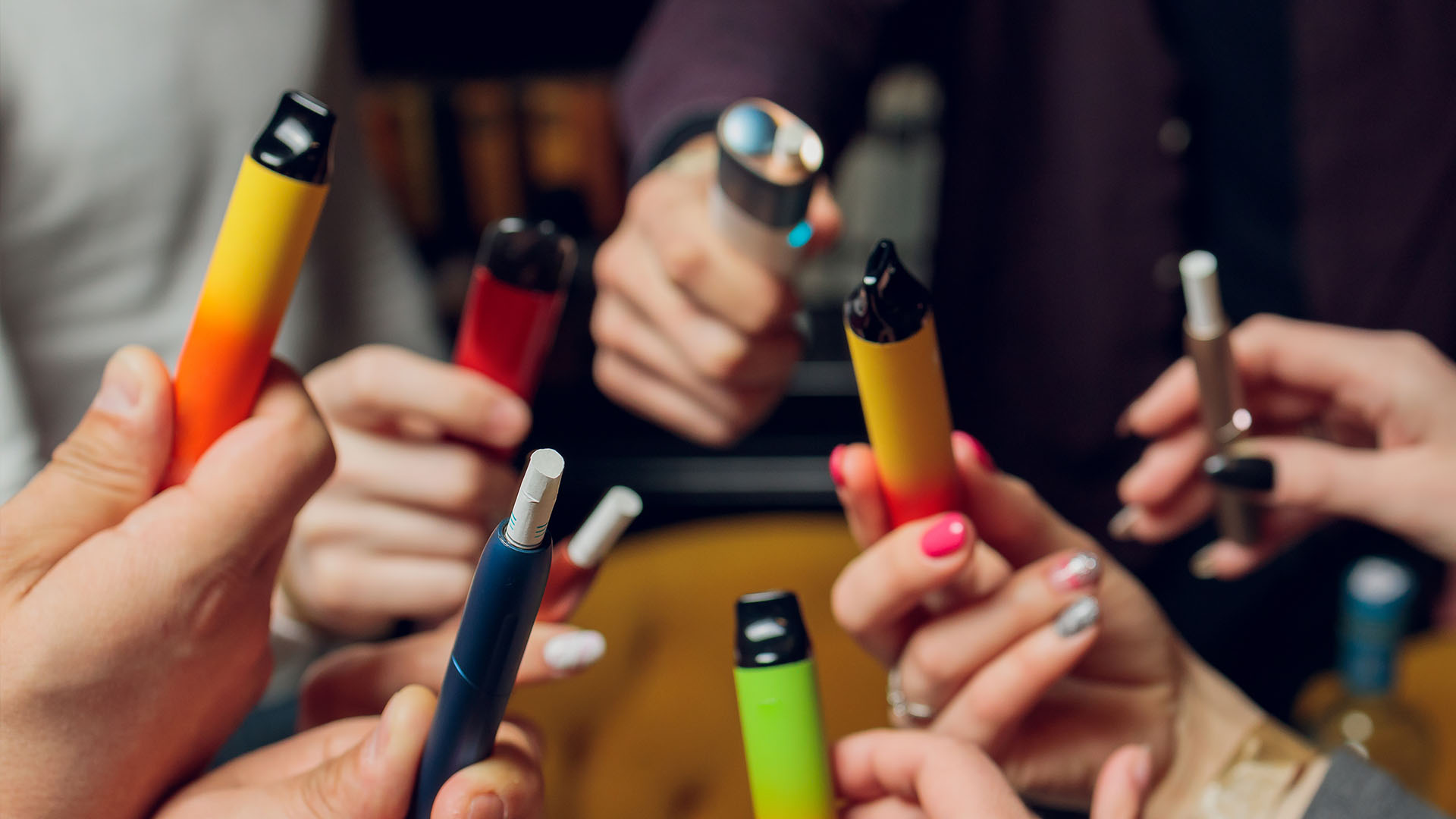 Hands holding various colourful vape pens in a social setting.