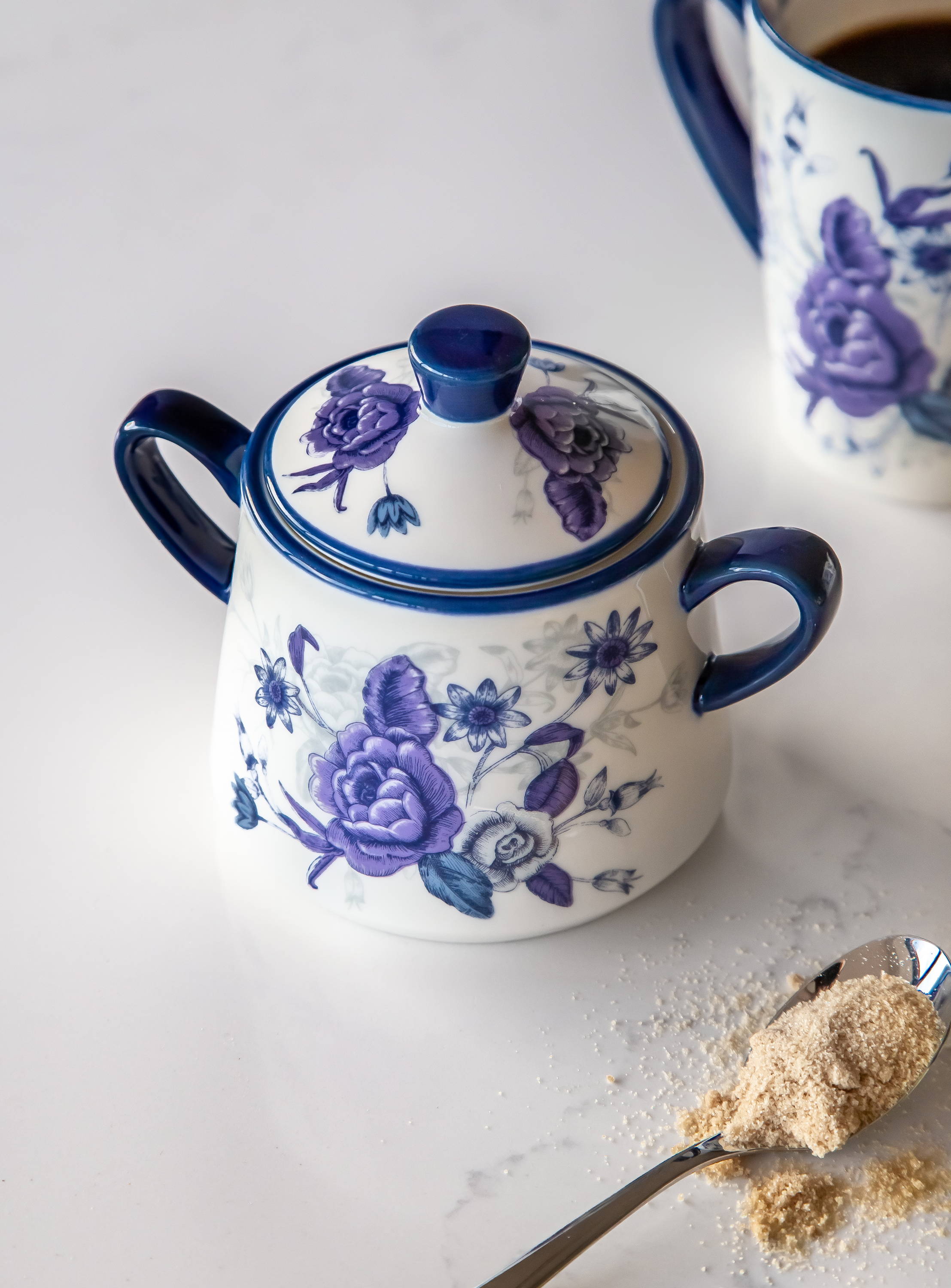 A blue floral sugar bowl with the lid on. There is a teaspoon with brown sugar on next to it.