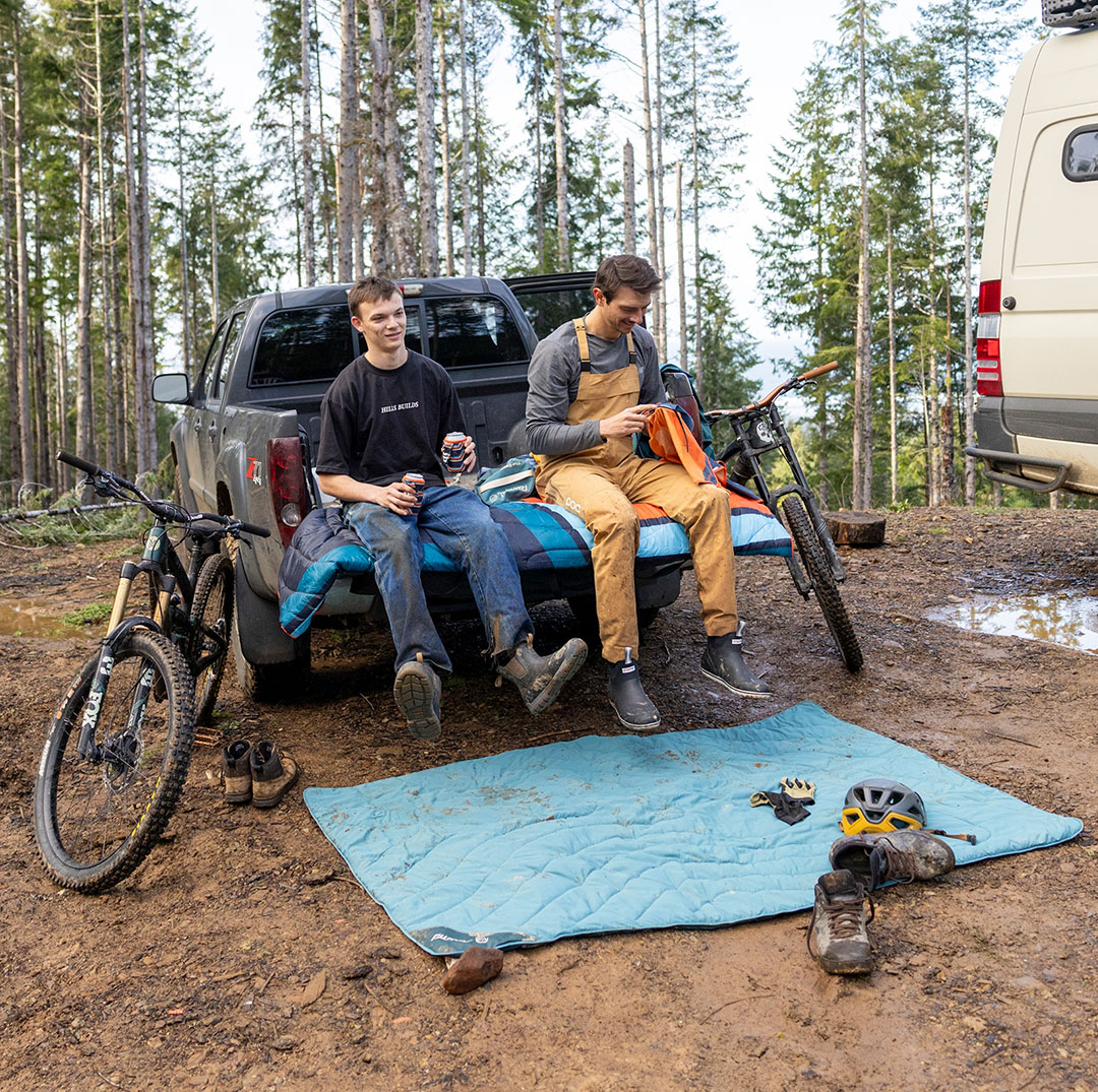 Two men sitting on a truck tailgate in a forested area.