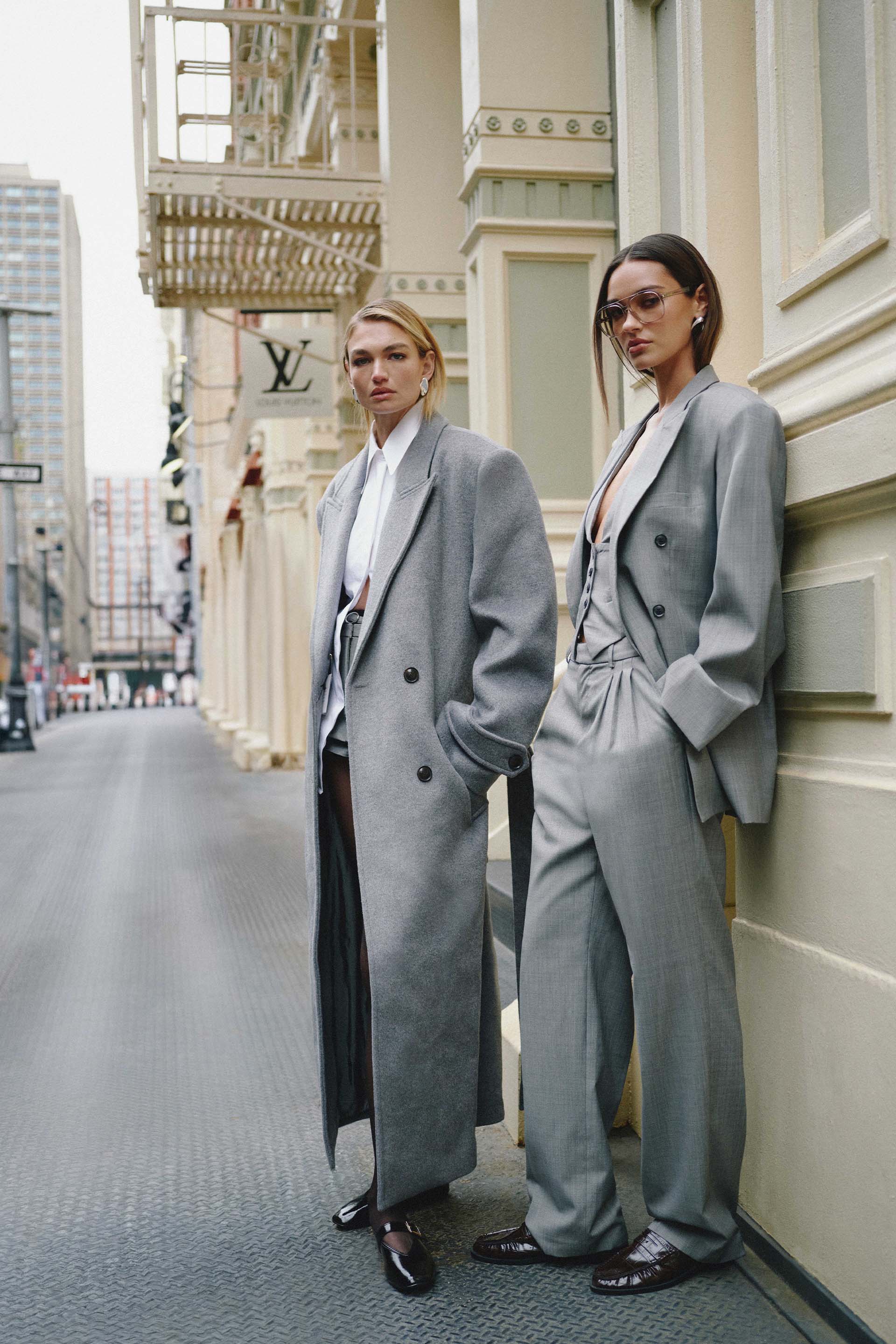 Two models wearing gray suits stand on a city street.