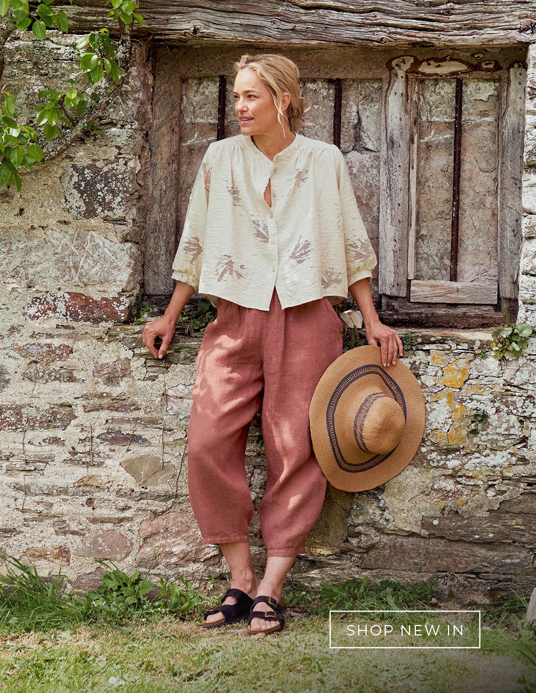 Woman wearing rust linen trousers and white textured shirt against a rustic wall 