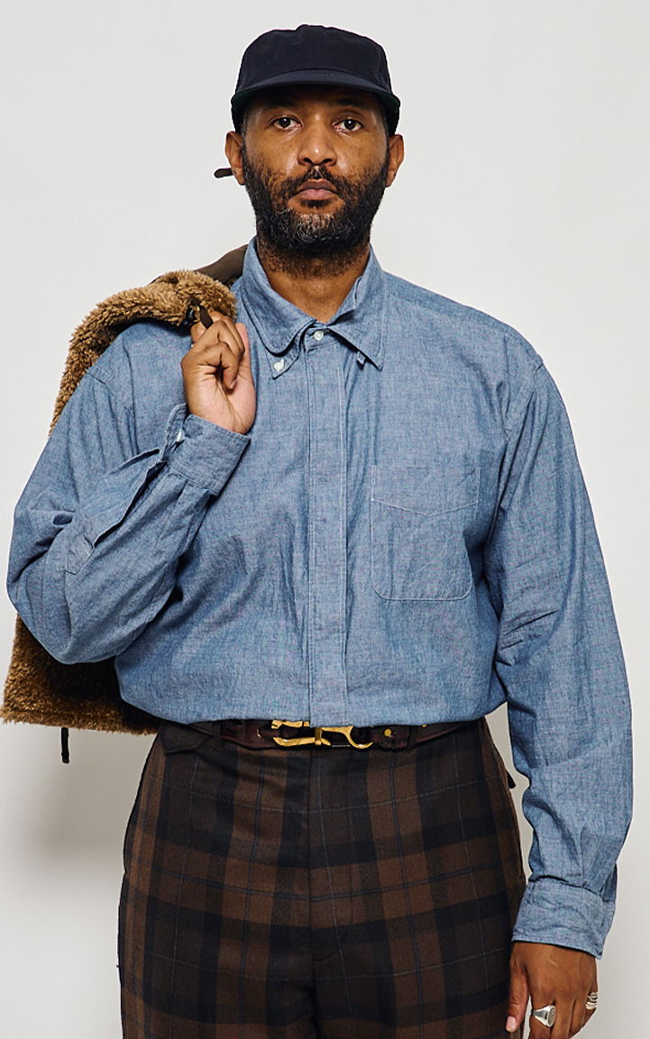 Man in a blue shirt holding a jacket, wearing a black cap and plaid pants.