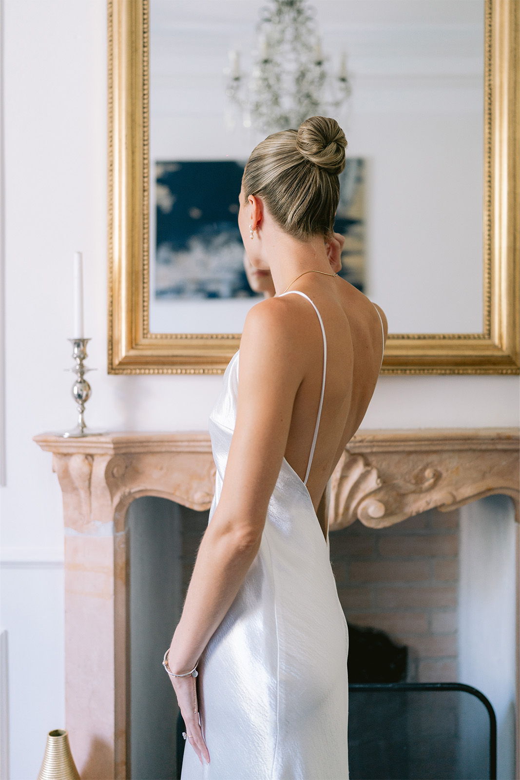 Woman in a sleek white dress standing by a mirror and fireplace.