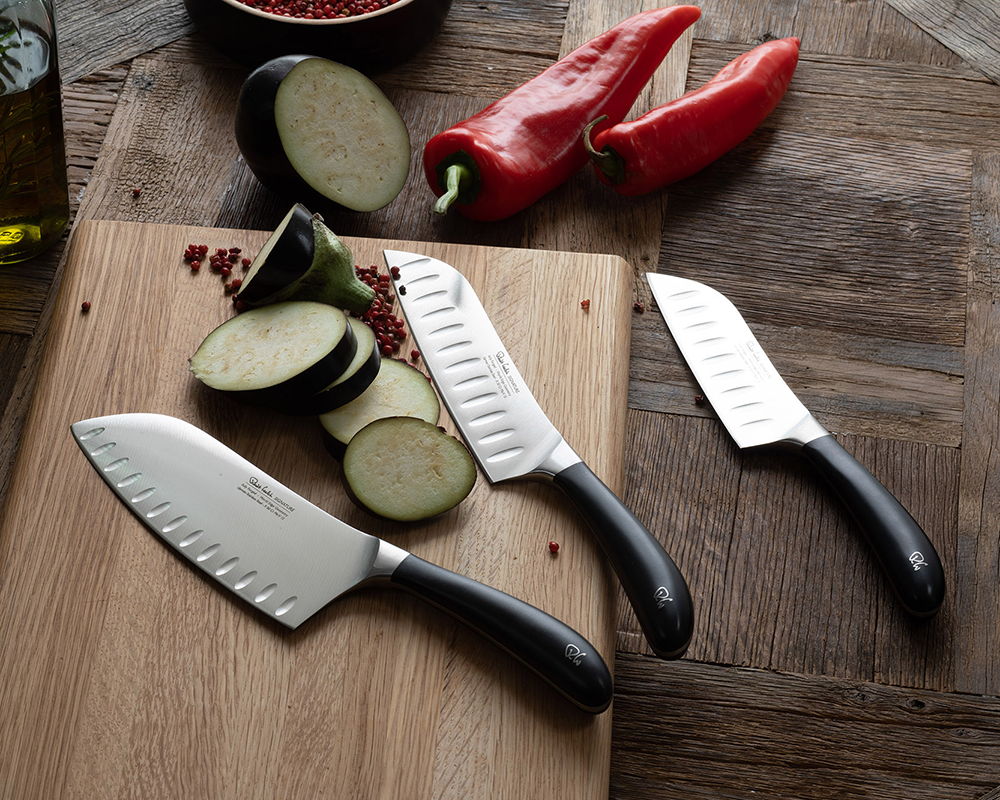 Signature Santoku Knives on a wooden board with sliced vegetables and red peppers.