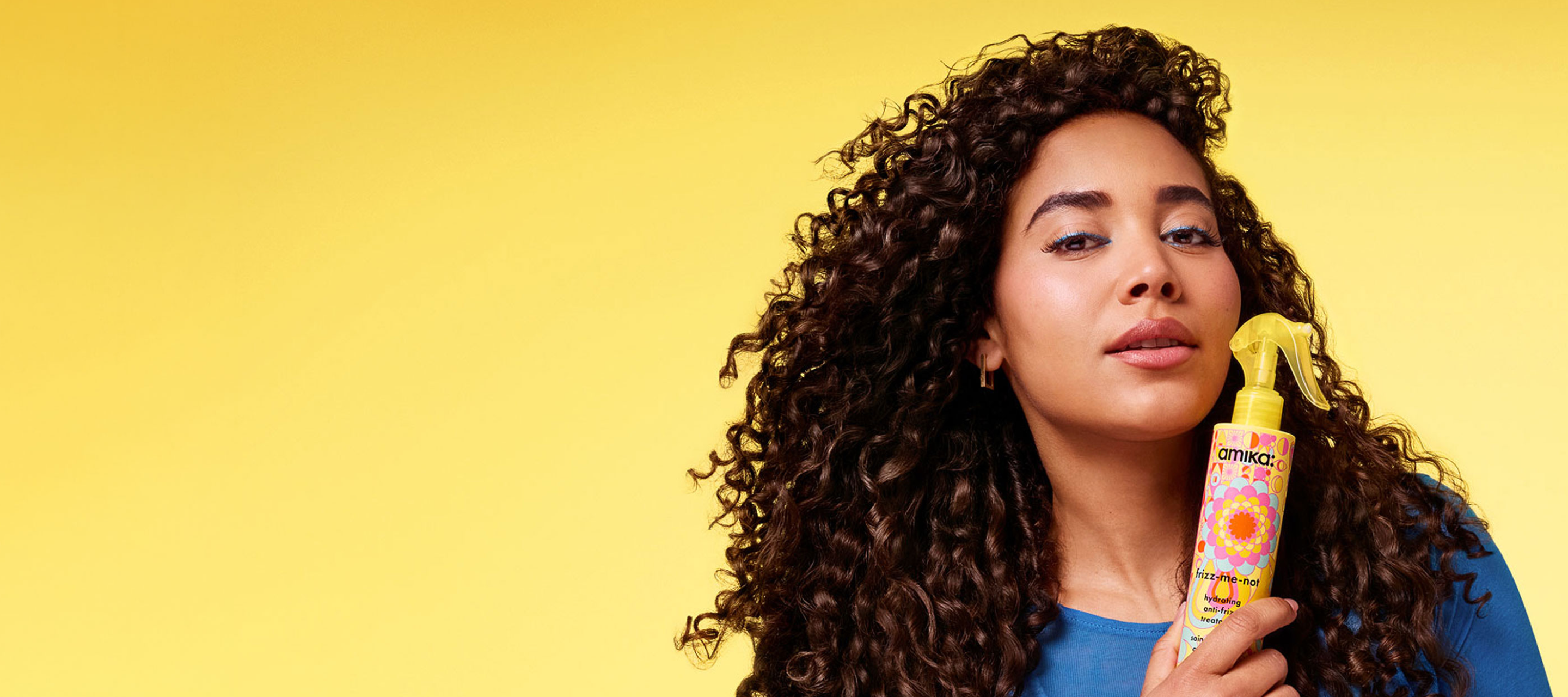 Woman with curly hair holding a hair product against a yellow background.