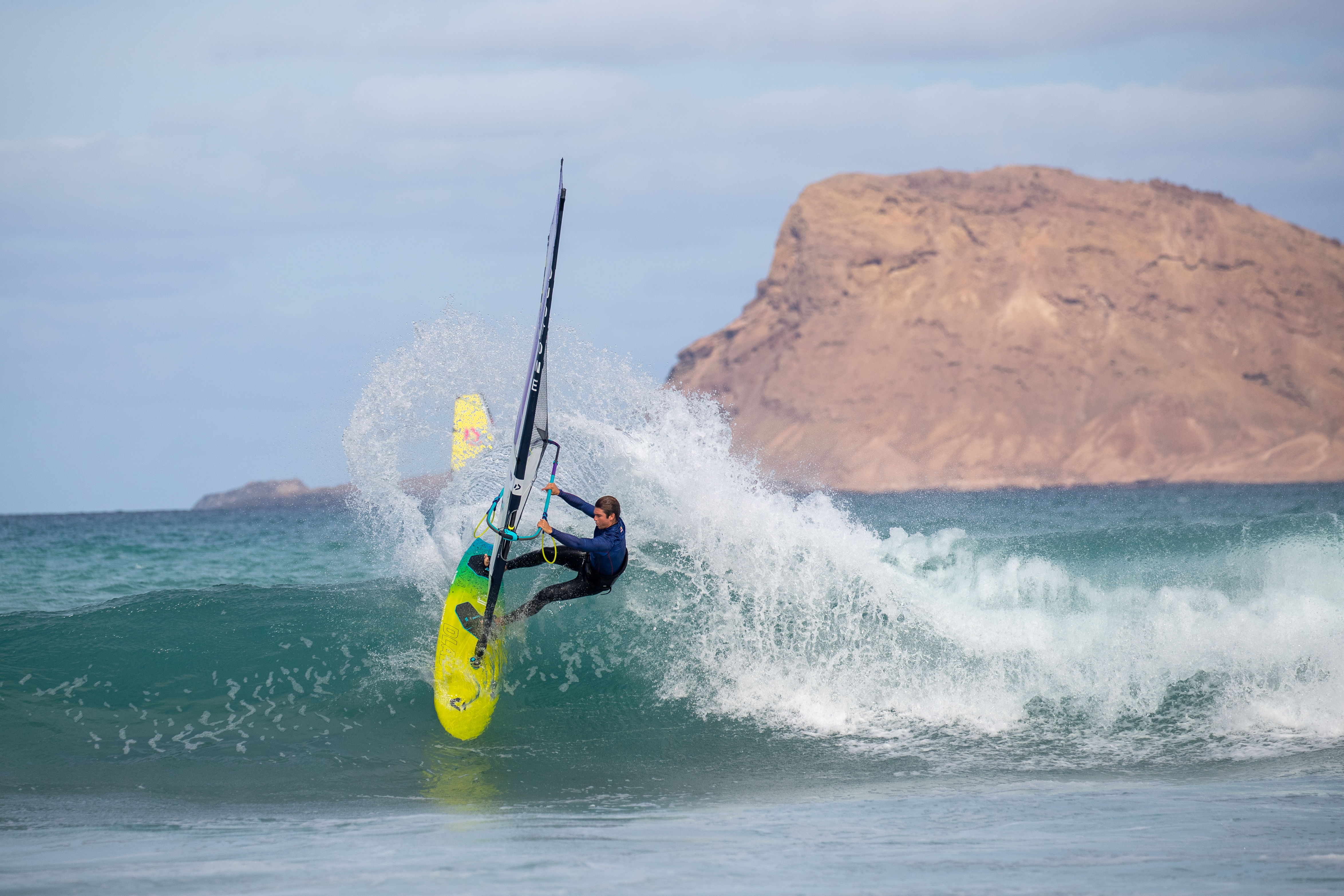 Surfer riding a wave with a yellow board near a rocky coastline.