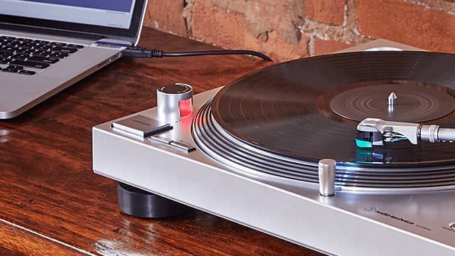 Turntable on a wooden surface next to a laptop against a brick wall.
