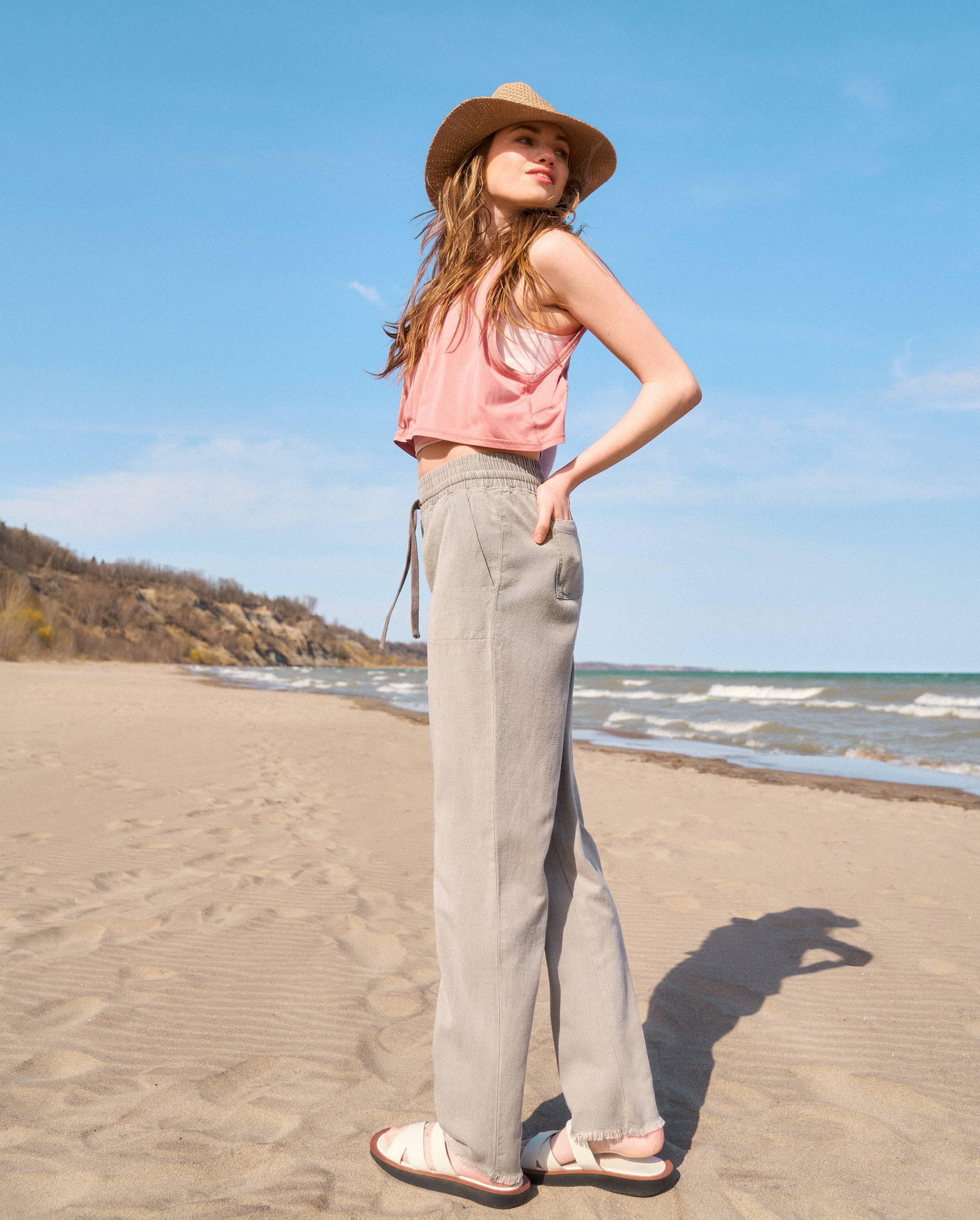 Tall woman standing on the beach wearing a pink tank top and taupe pants 