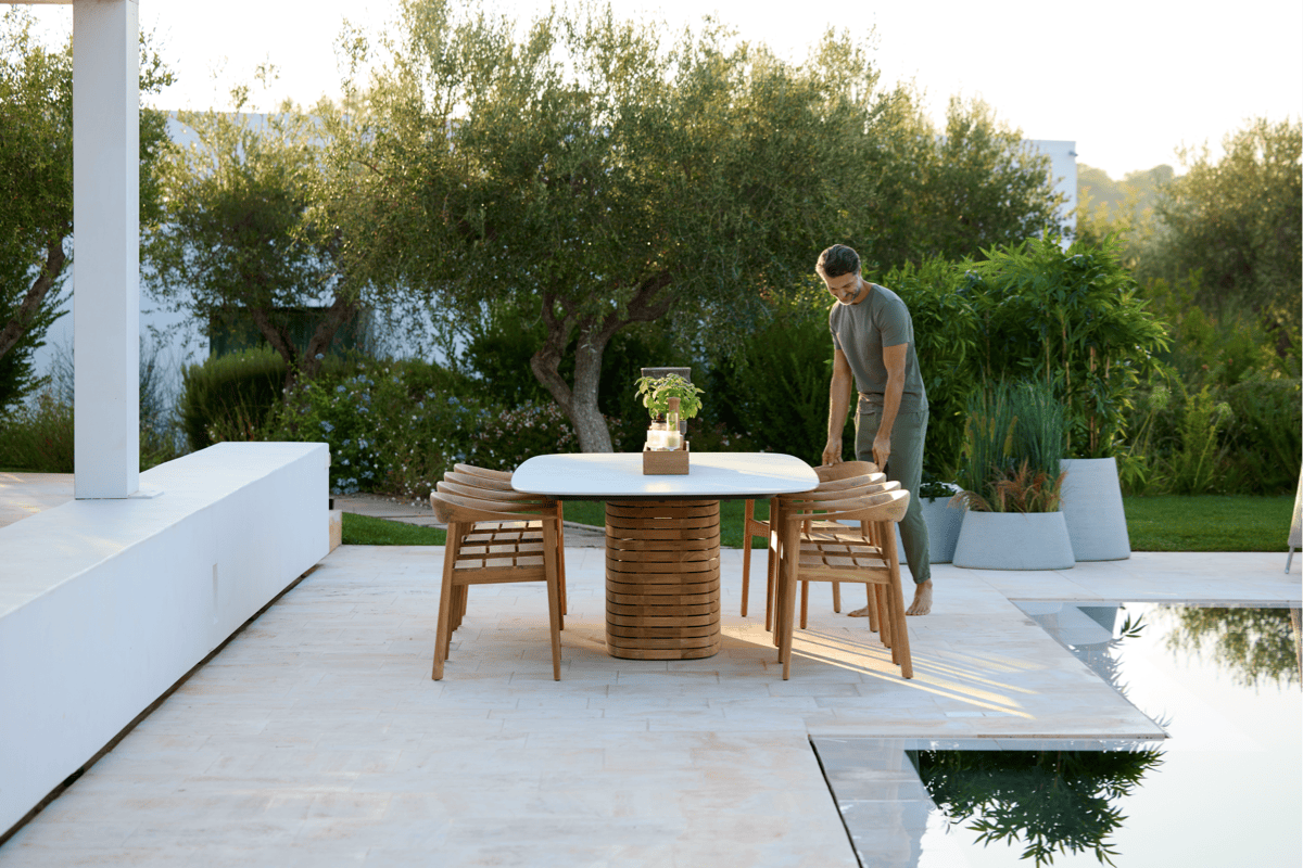 Person arranging wooden dining chairs around a round outdoor table on a modern patio beside a pool, with lush greenery in the background.