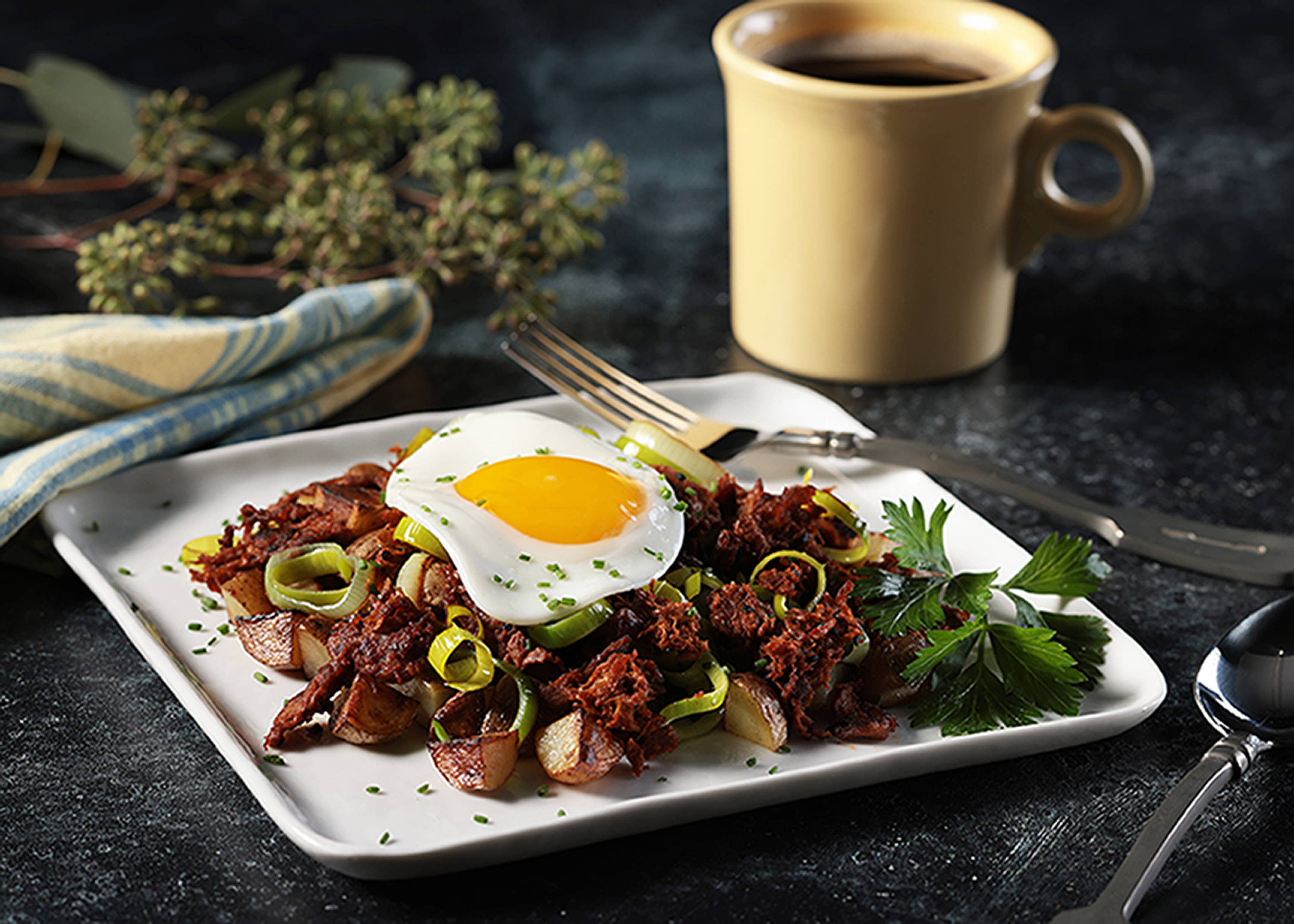 Texas BBQ Harvest Shreds hash with new potatoes and leeks, and topped with a fried egg, on a square white plate with a fork. Folded yellow and blue napkin and a yellow mug of coffee.