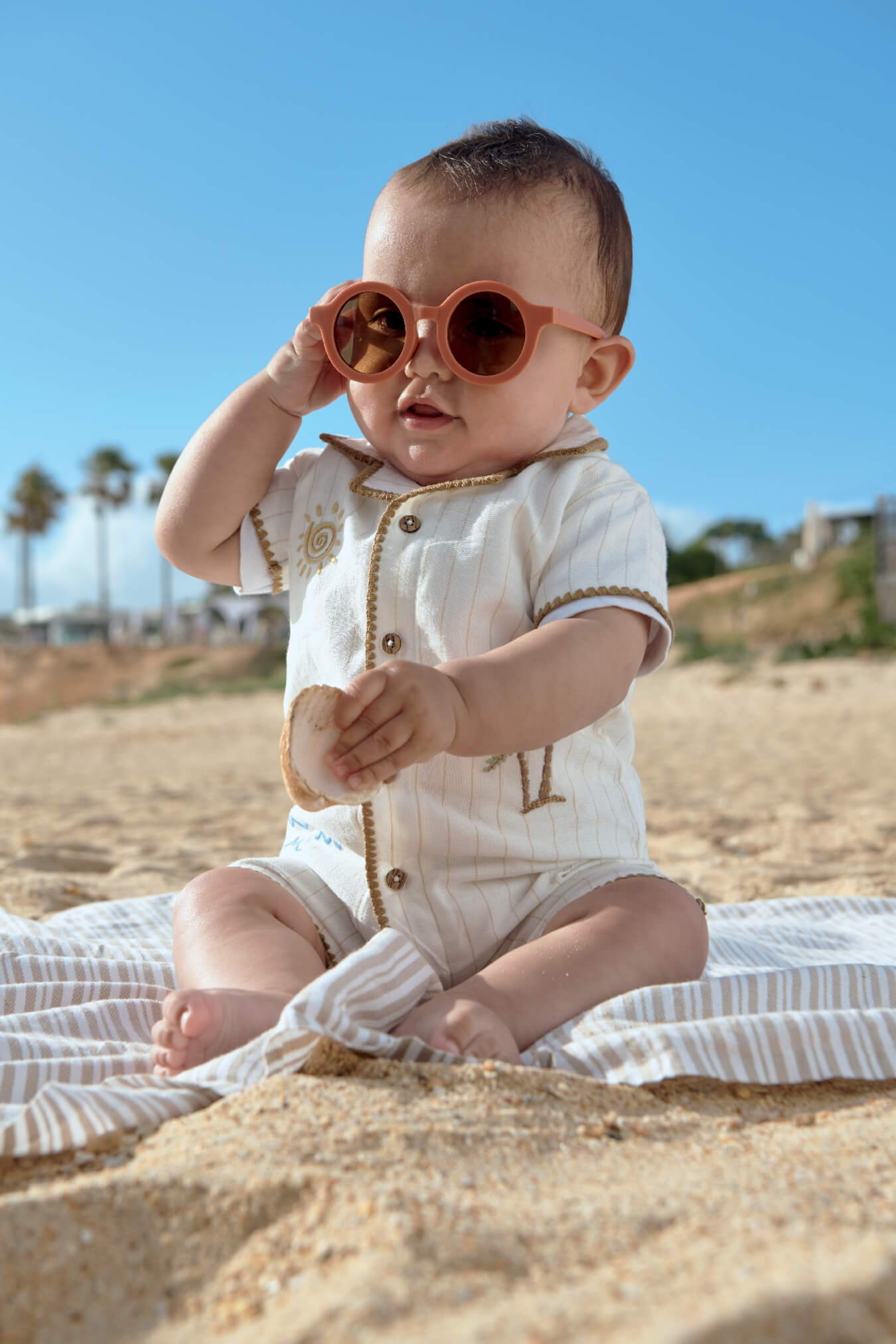 Baby wearing sunglasses, sitting on a beach towel in the sand.