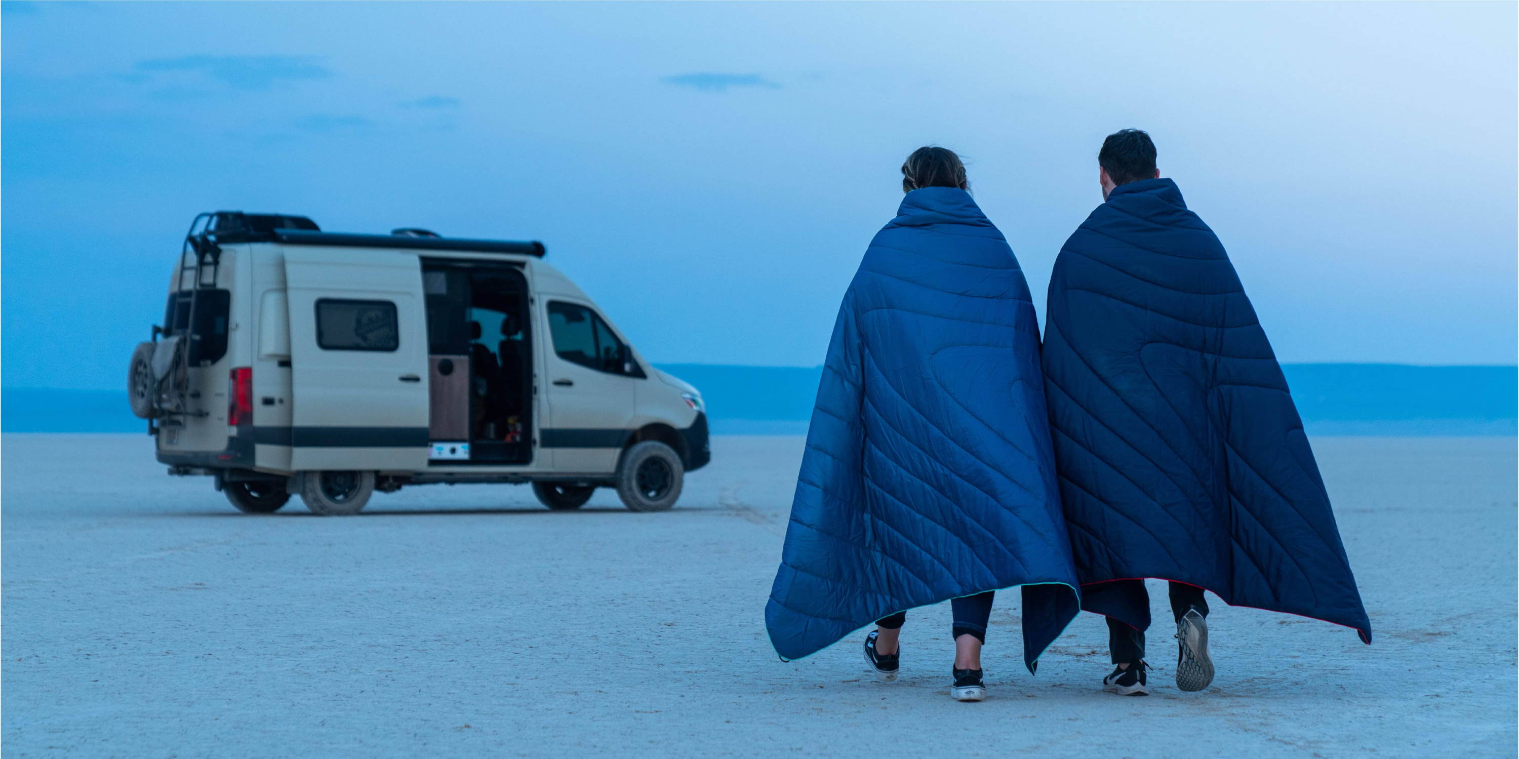Man and Woman Walking Through Desert Towards Camper Van While Wrapped In Original Puffy Blankets