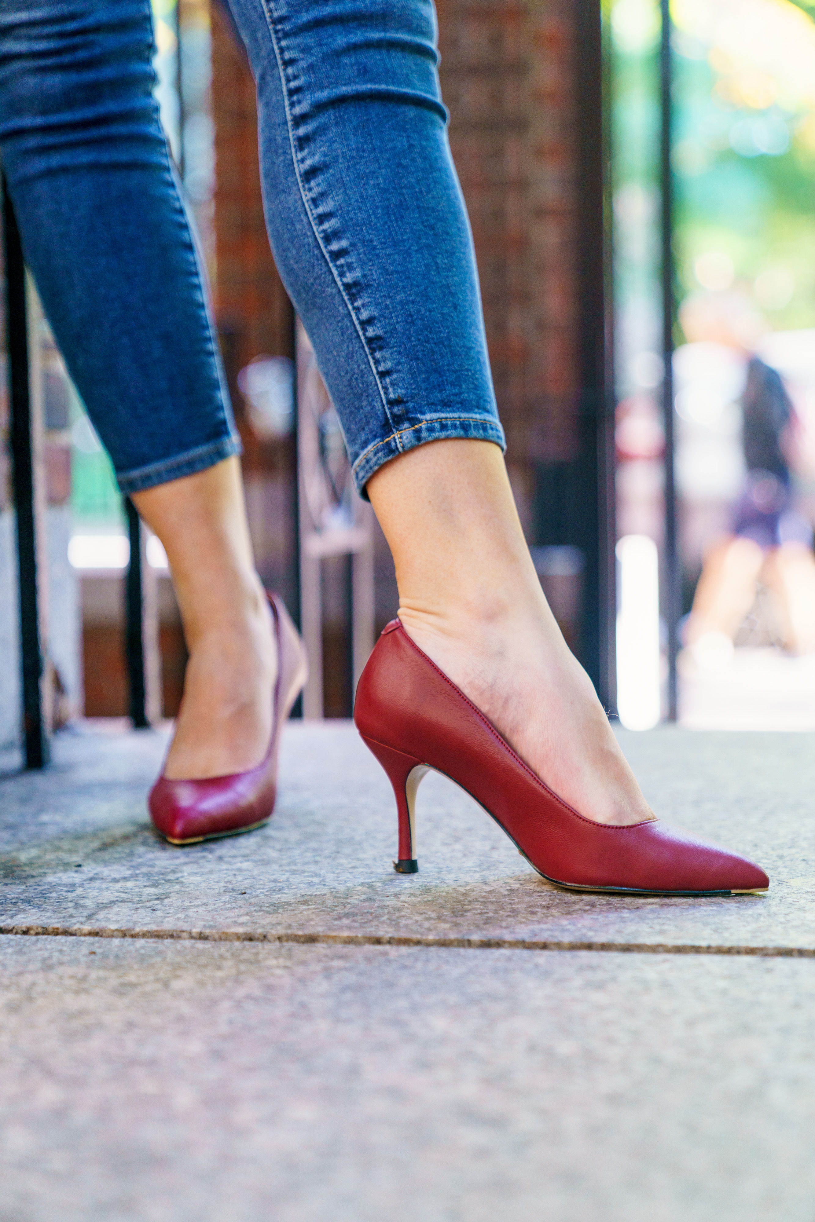 A person wearing red high heels and blue jeans walking on pavement.