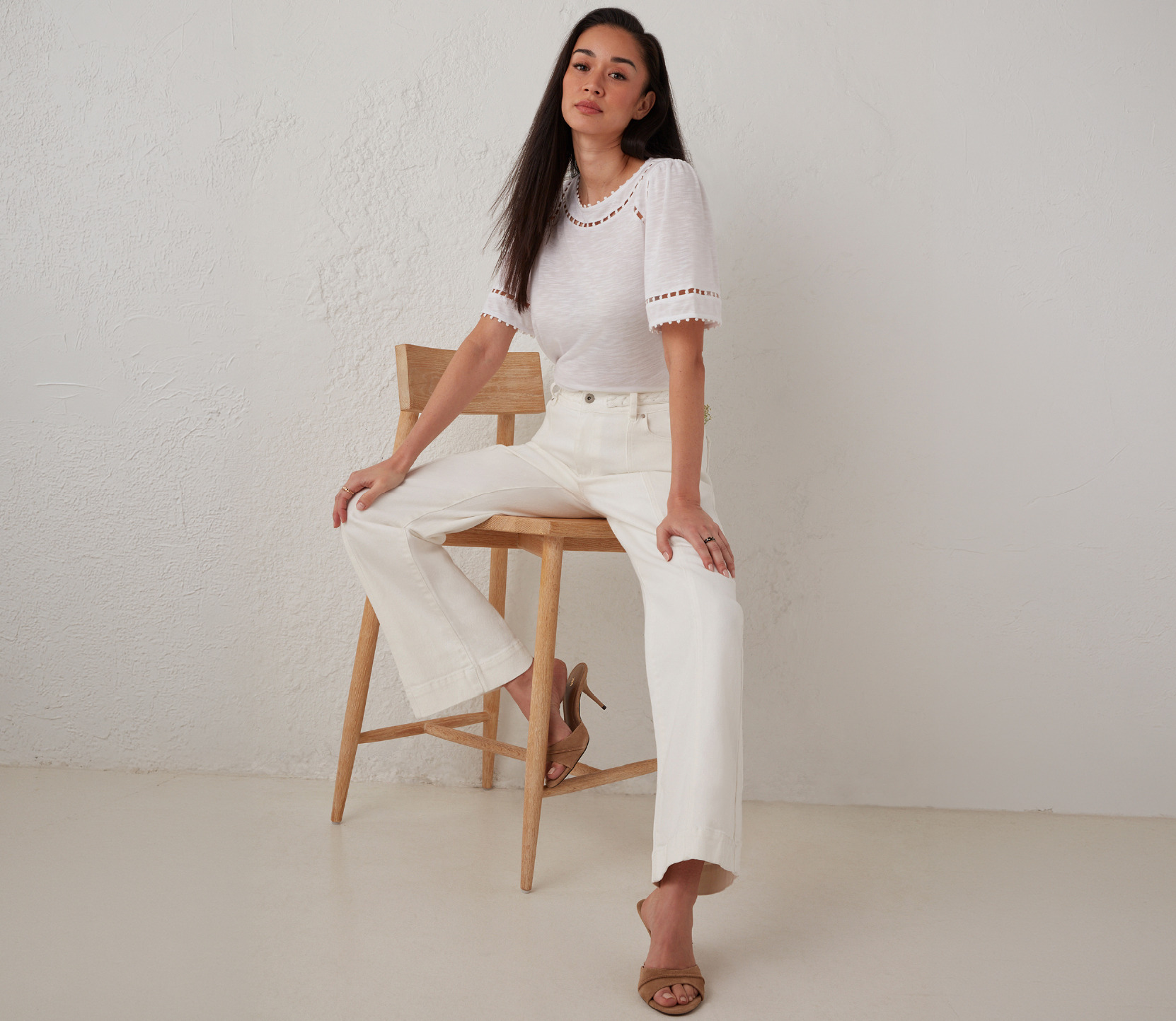 Woman sitting on a stool, wearing a white top and light-colored pants.