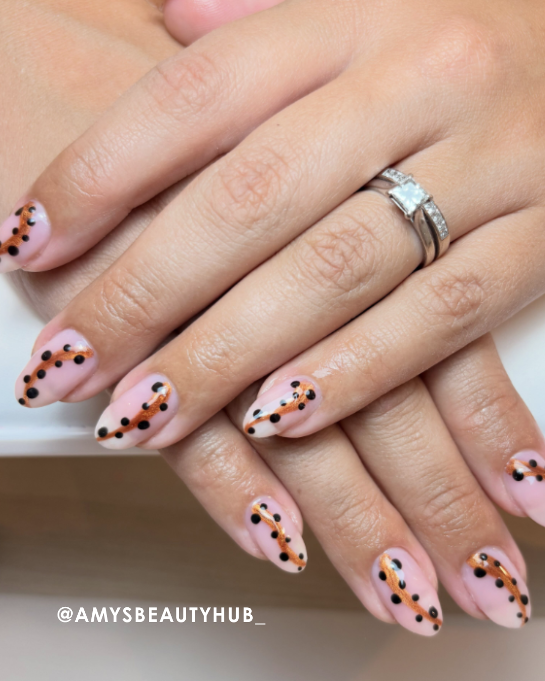 Hands with manicured nails featuring pink polish and black dots, wearing a ring.