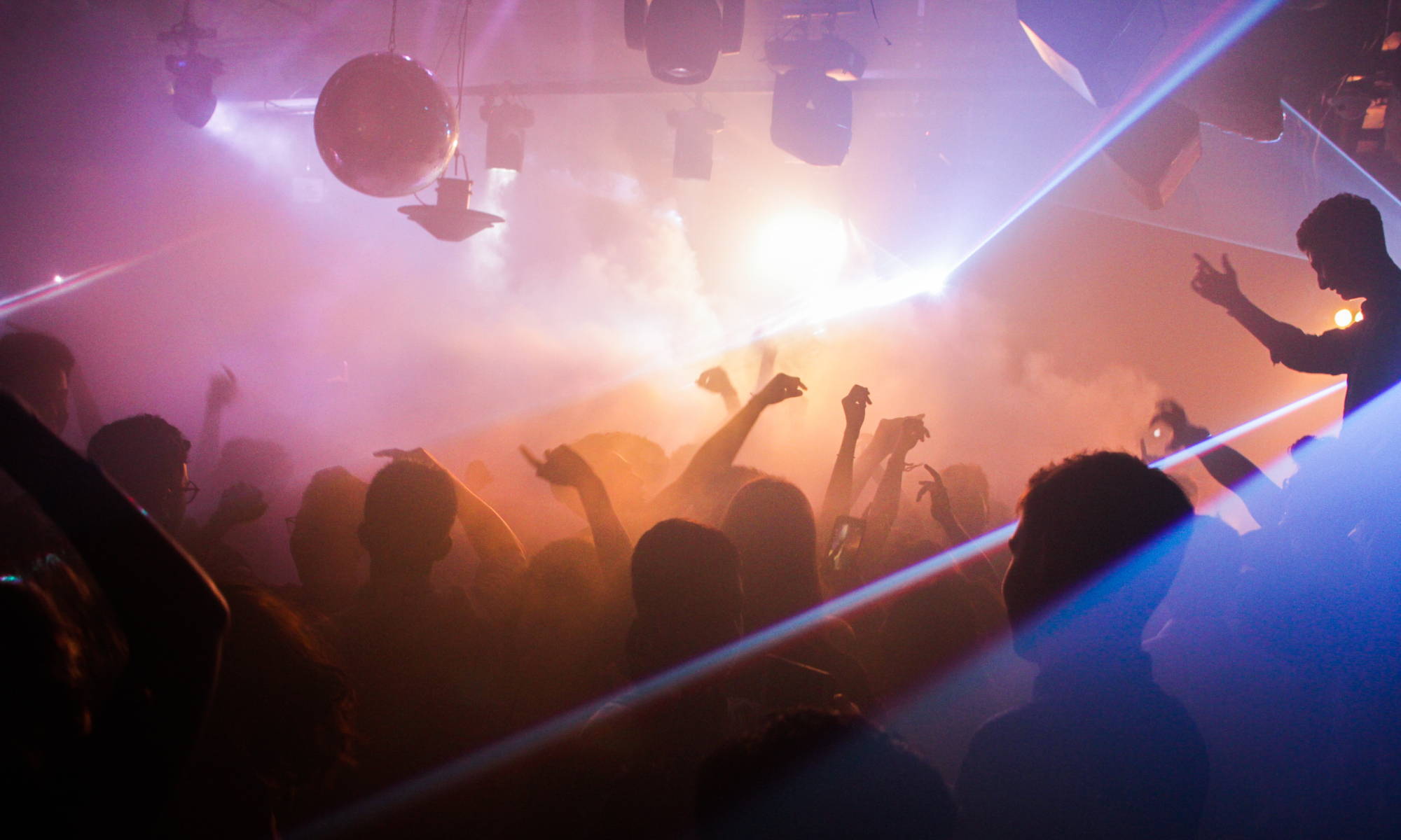 A crowd dances with their hands in the air at a busy nightclub with fog and flashing lights. Image by Sarthak Navjivan. 