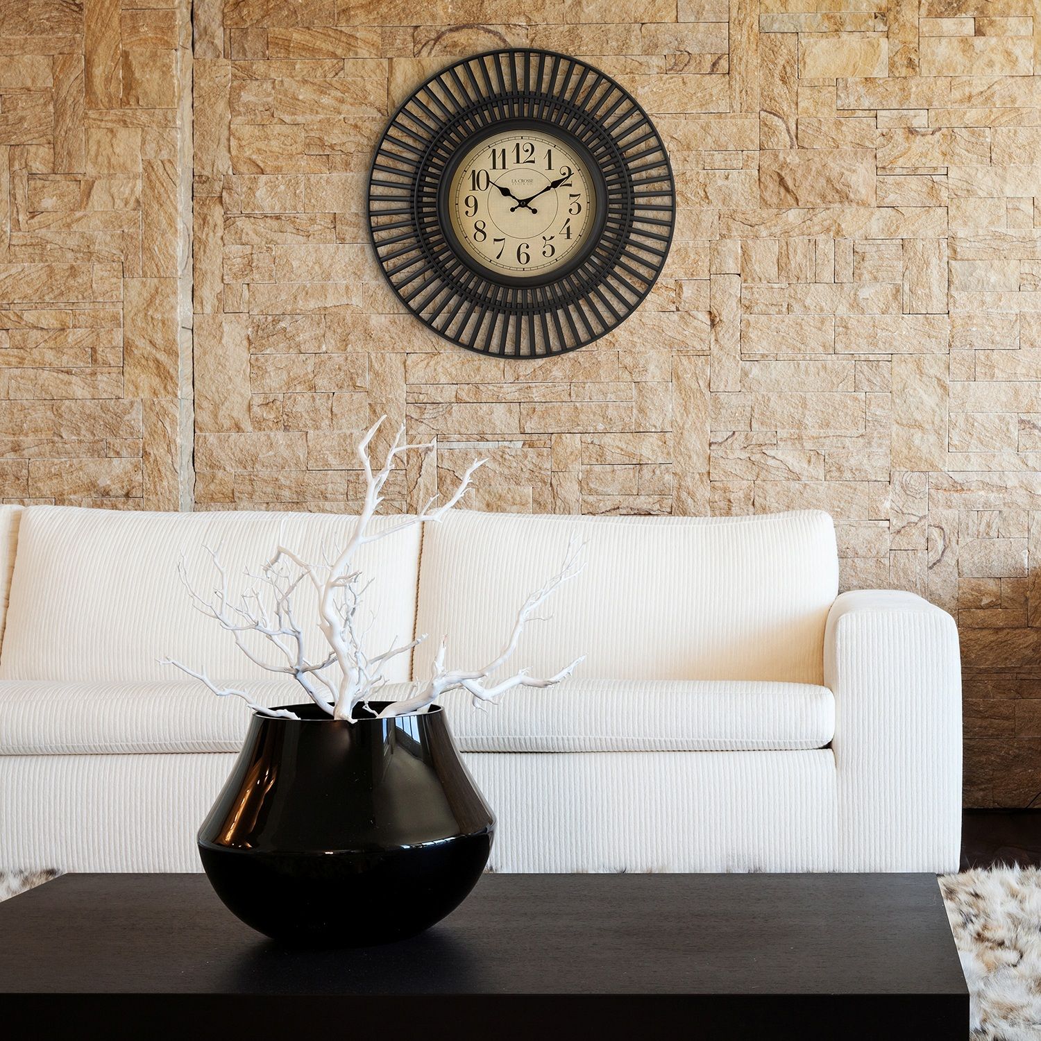 20-inch Covington Wall Clock hanging on a stone wall in a modern living room above a cream couch in front of a coffee table with a decorative vase. 