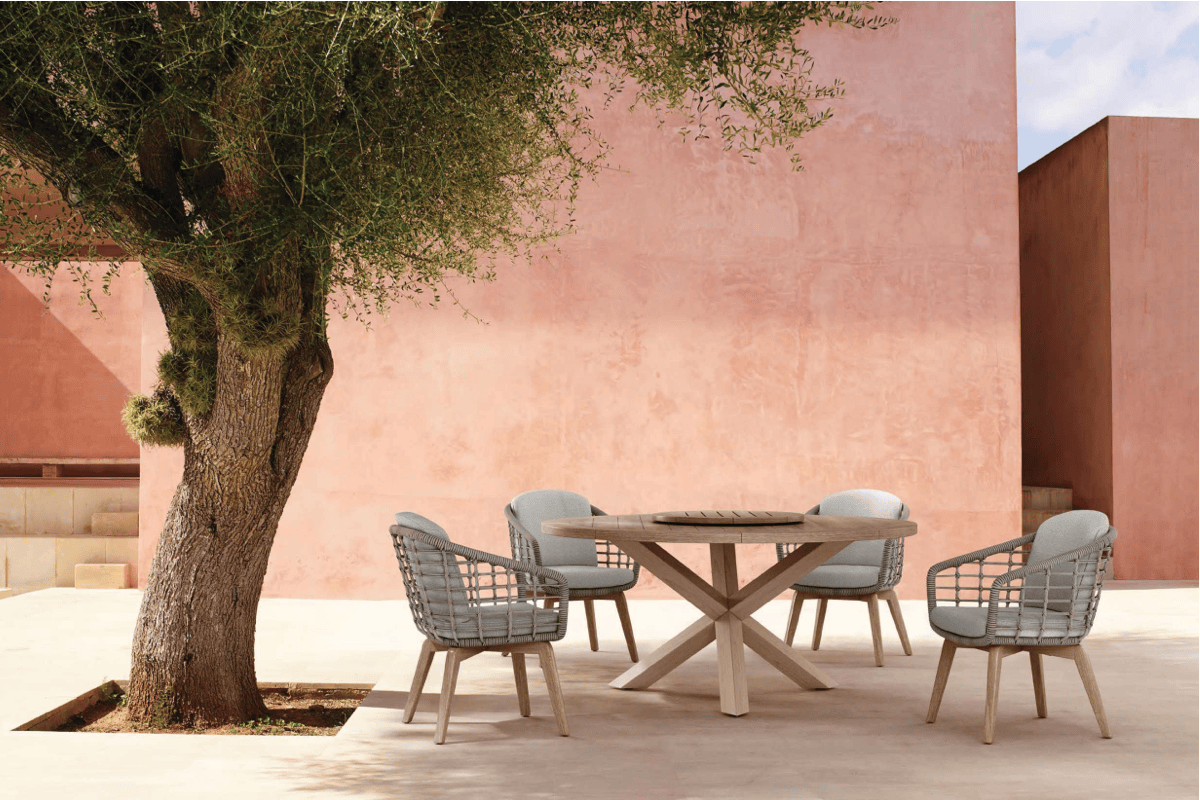 Round pedestal outdoor dining table with woven dining chairs set beside a blush stucco wall and mature olive tree in a serene courtyard.