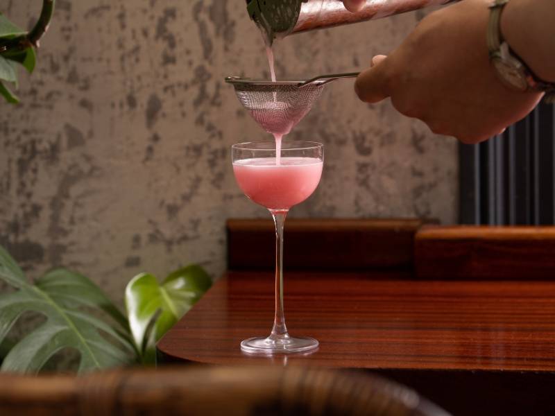Strawberry and lychee margarita being strained into a cocktail glass on a wooden surface 