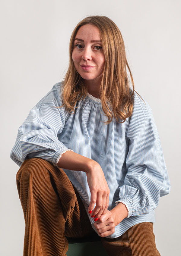 Tanya sitting, wearing a blue blouse and brown pants, smiling at the camera.
