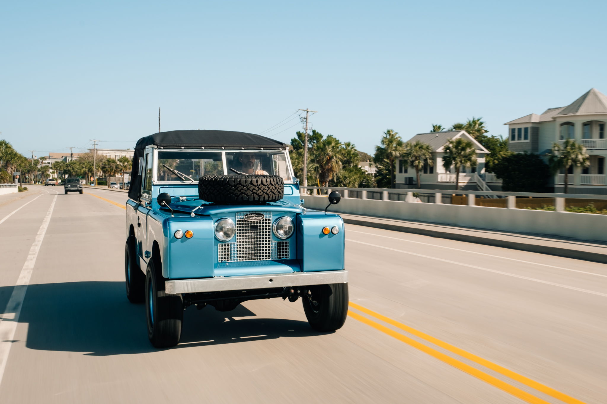 Blue vintage vehicle driving on a sunny road with houses nearby.