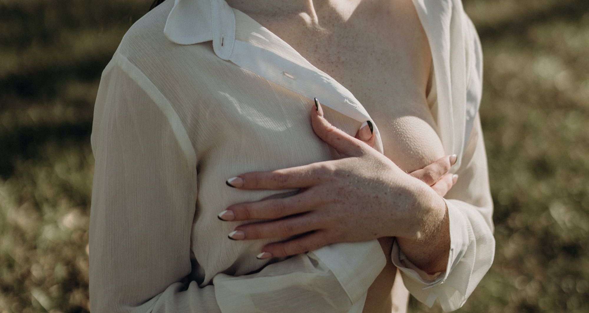  A woman wearing a lightweight, unbuttoned, white blouse stands outside and covers her breasts with both hands