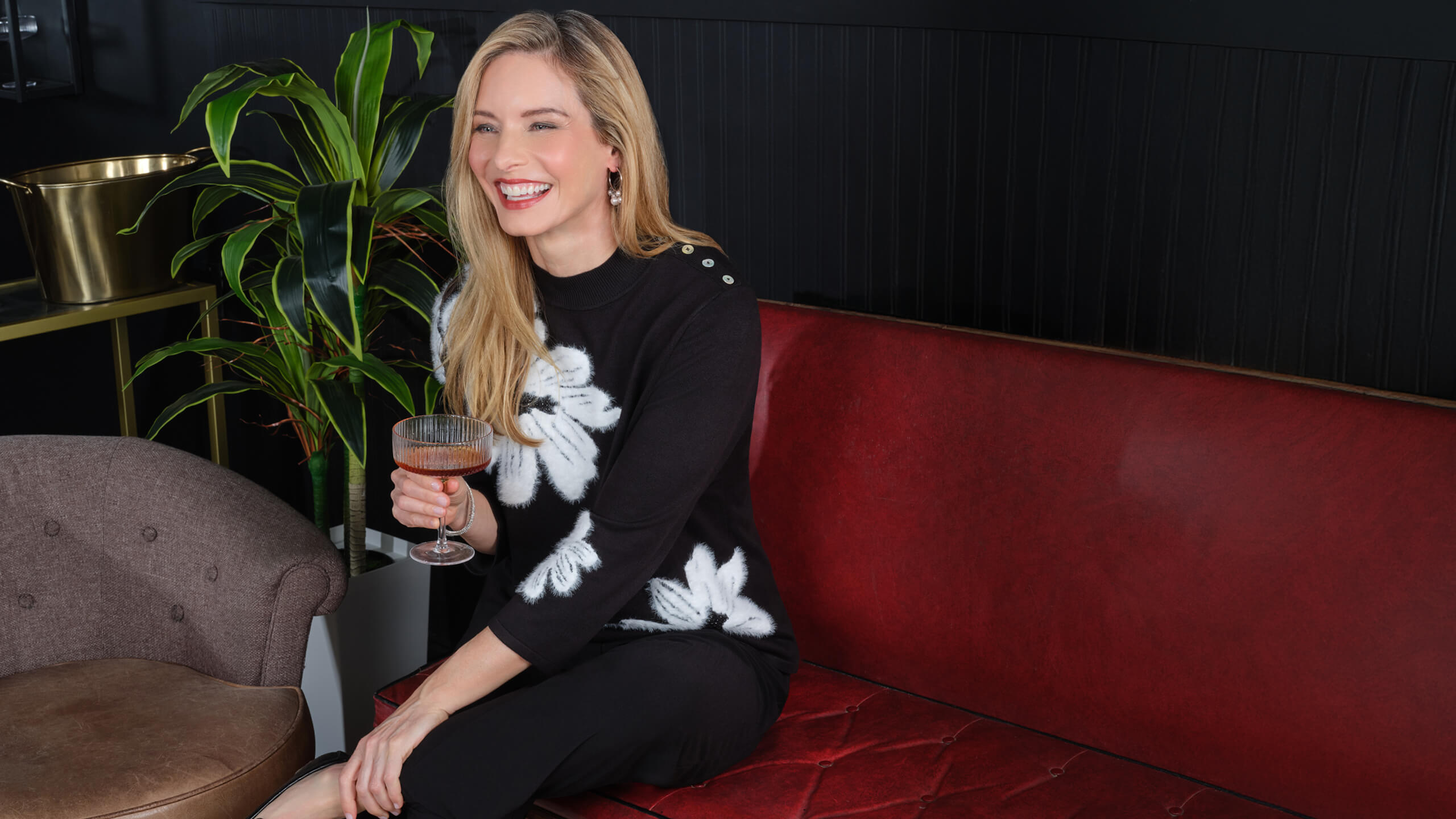Smiling woman in a floral sweater sitting on a couch with a drink.