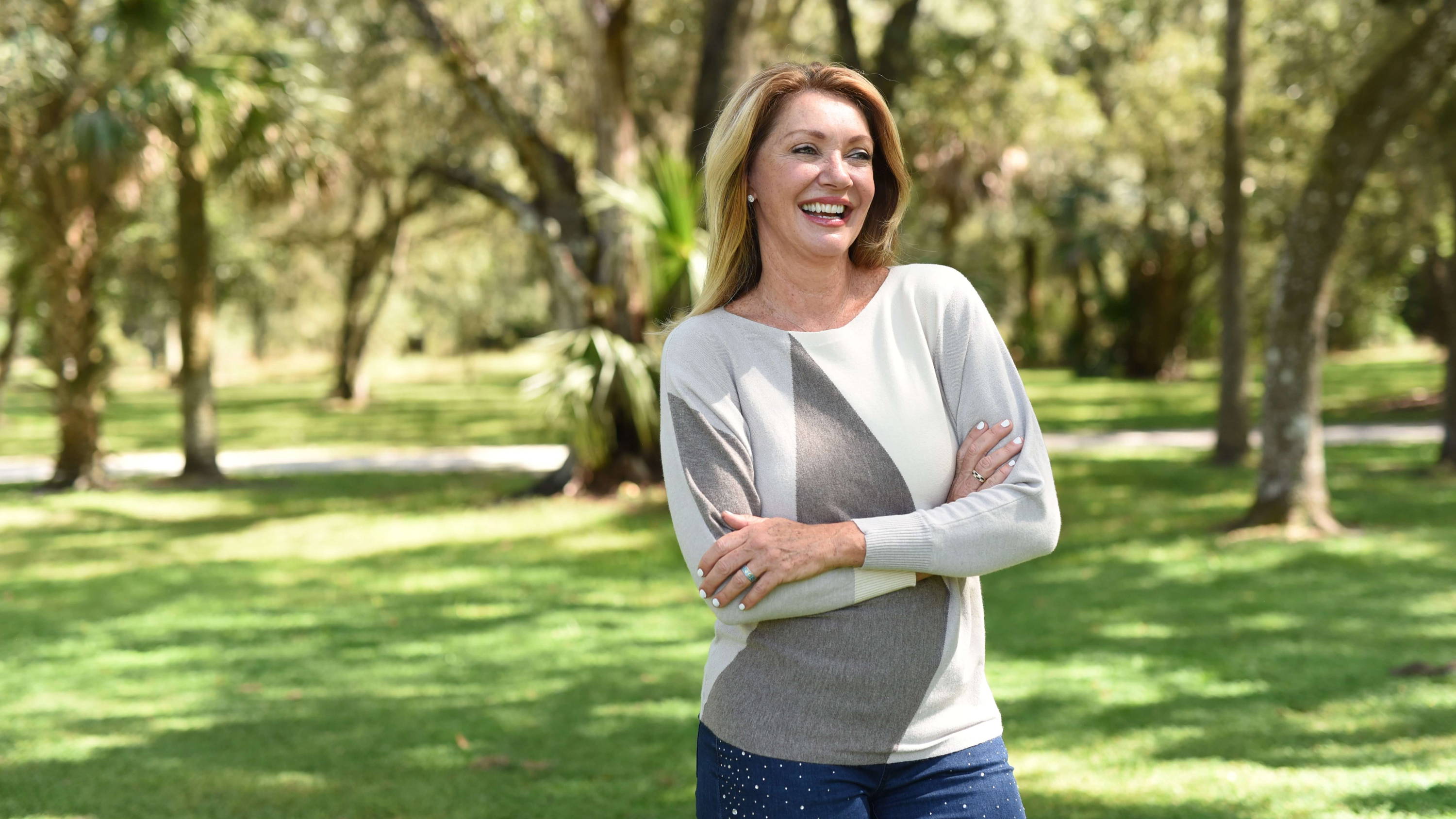 A blonde woman laughs as she stands with her arms crossed in a park. 