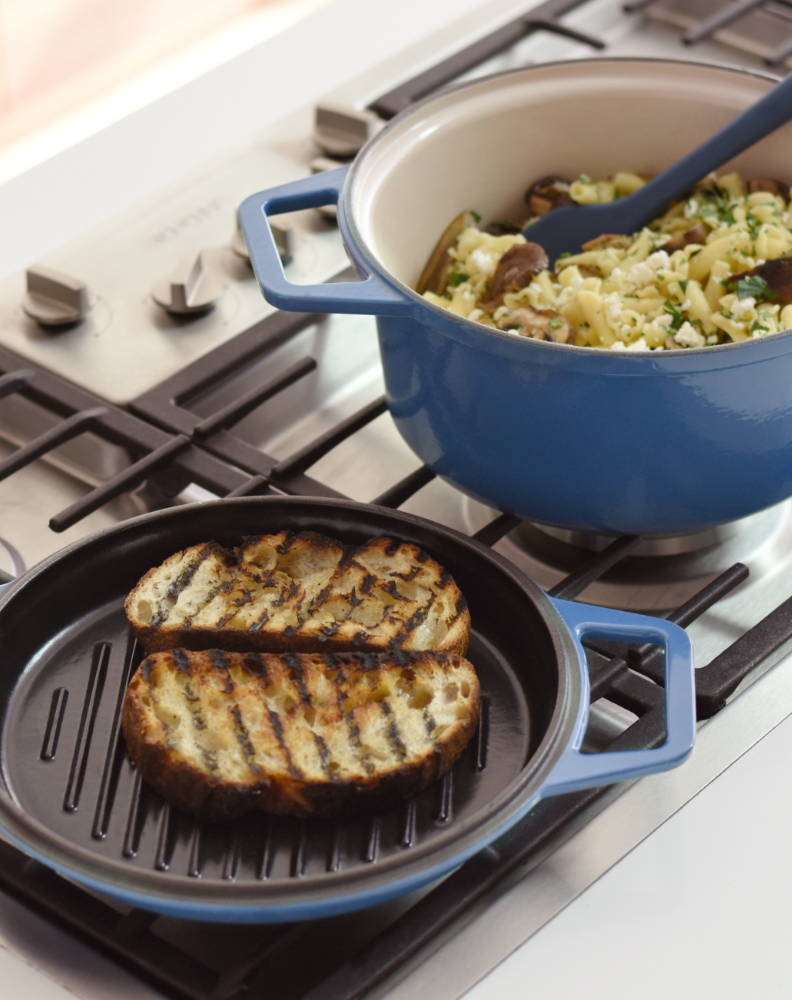 A blue Misen Grill Lid toasting buttered bread on a stovetop next to a blue Misen Dutch Oven full of sausage and pasta.