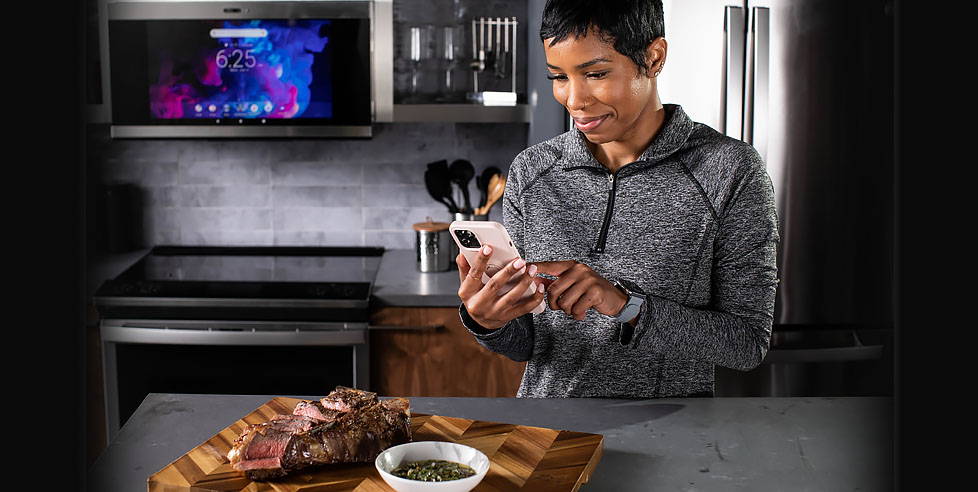 Woman checking her smartphone with steak on the island and GE Profile smart appliances in the background