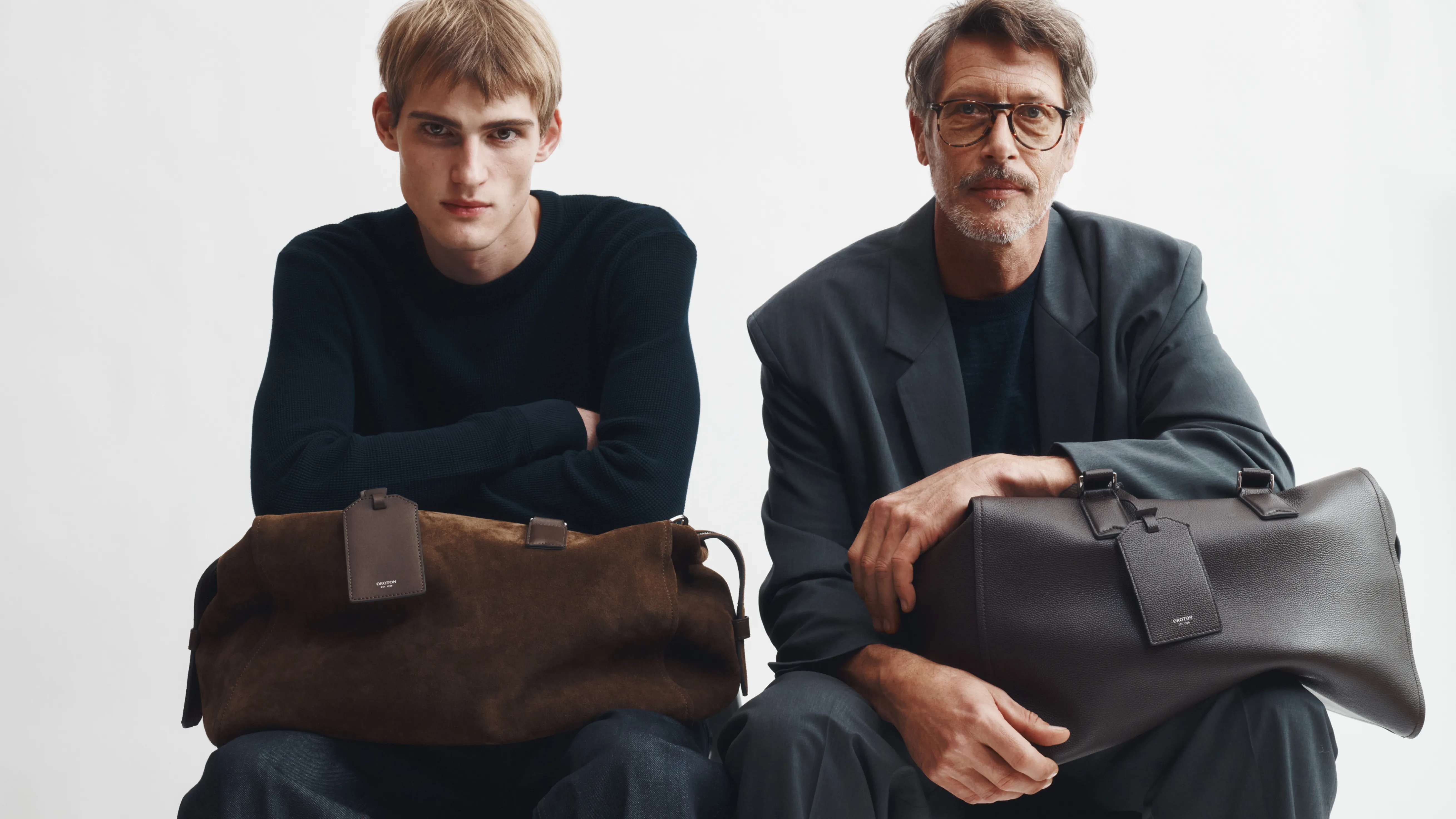 Two men sitting with bags, one older and one younger, on a plain background.