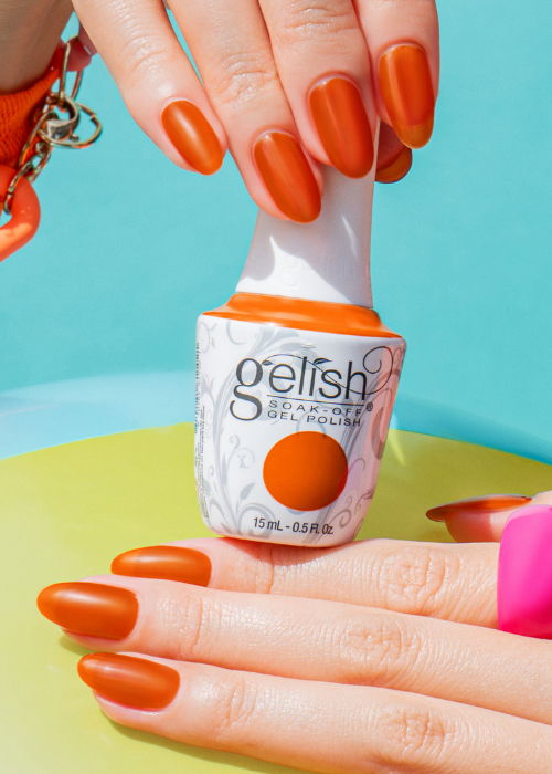 Orange nail polish bottle with manicured hands in a colorful setting.