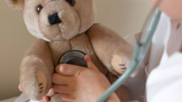 Ein Kind in einem weißen Kittel spielt Doktor. Es nutzt ein Stethoskop, um einen Teddy zu untersuchen.