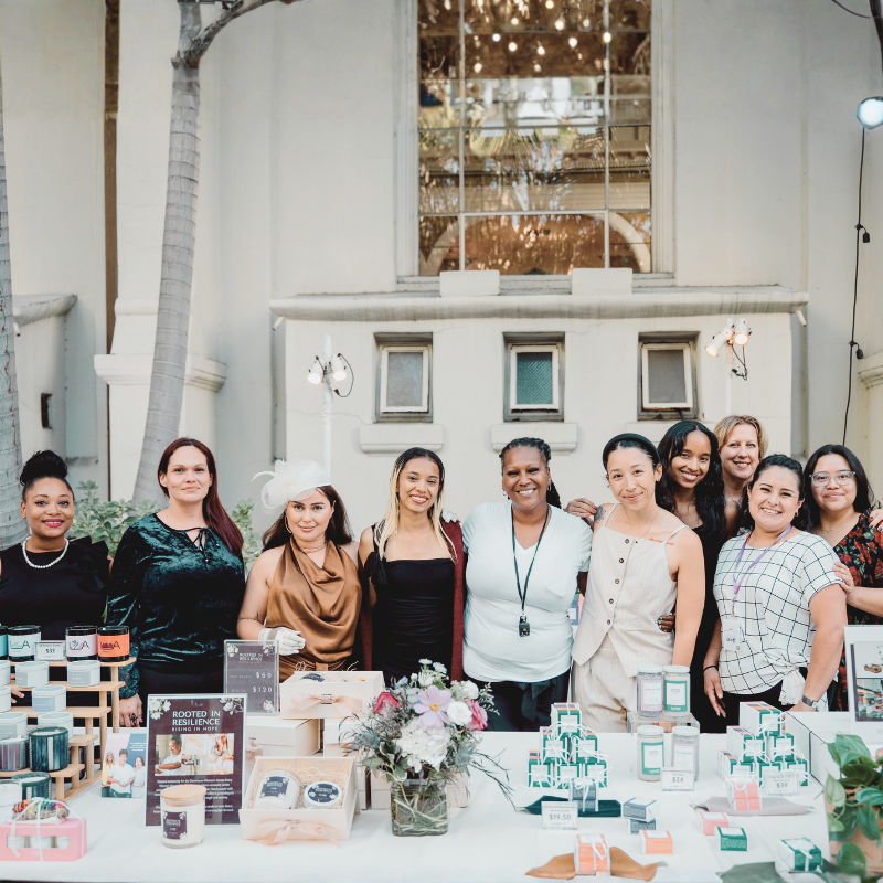 Group of women posing at a market stall with various products displayed.