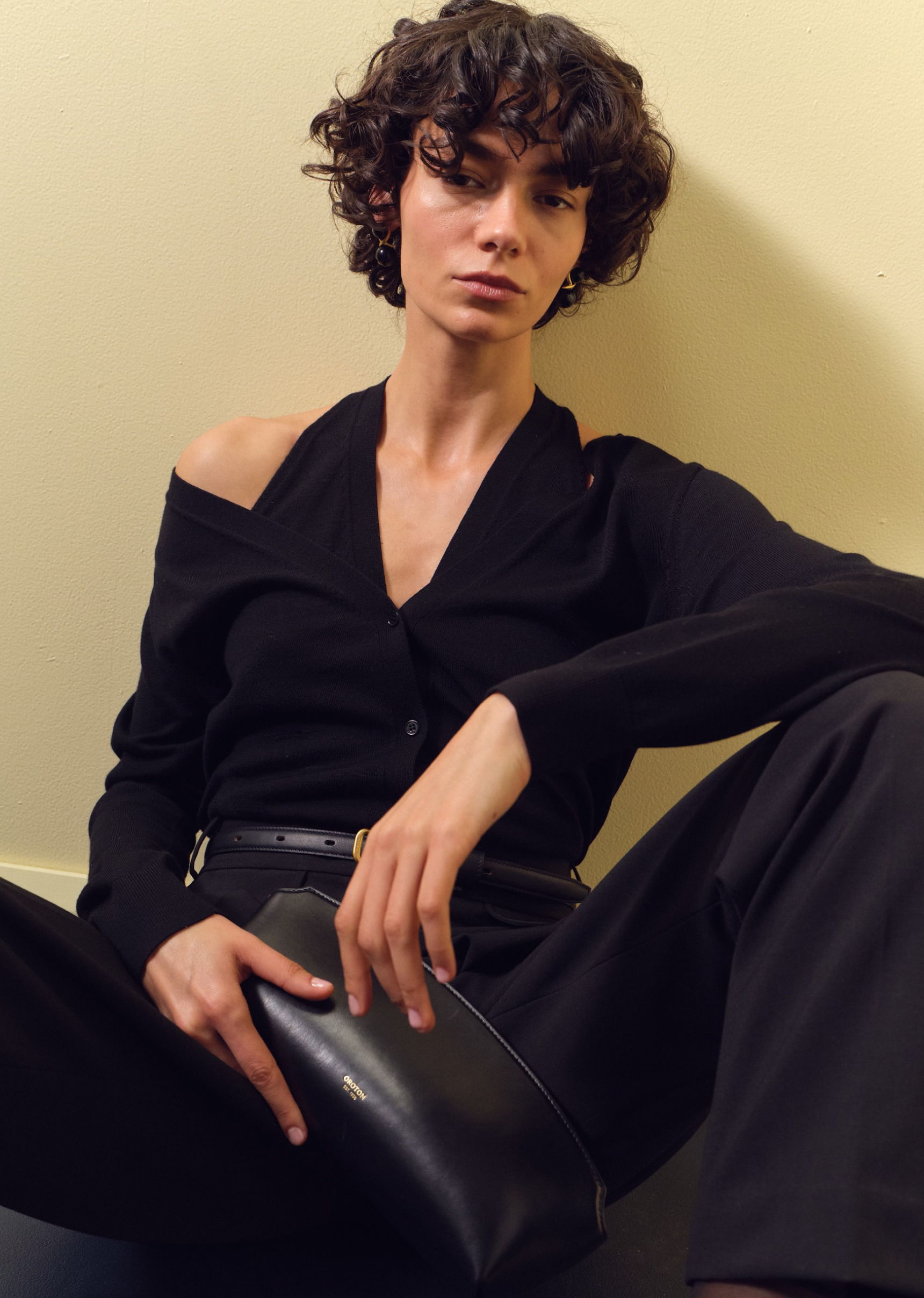 Person sitting against a wall, wearing a black outfit and holding a clutch.