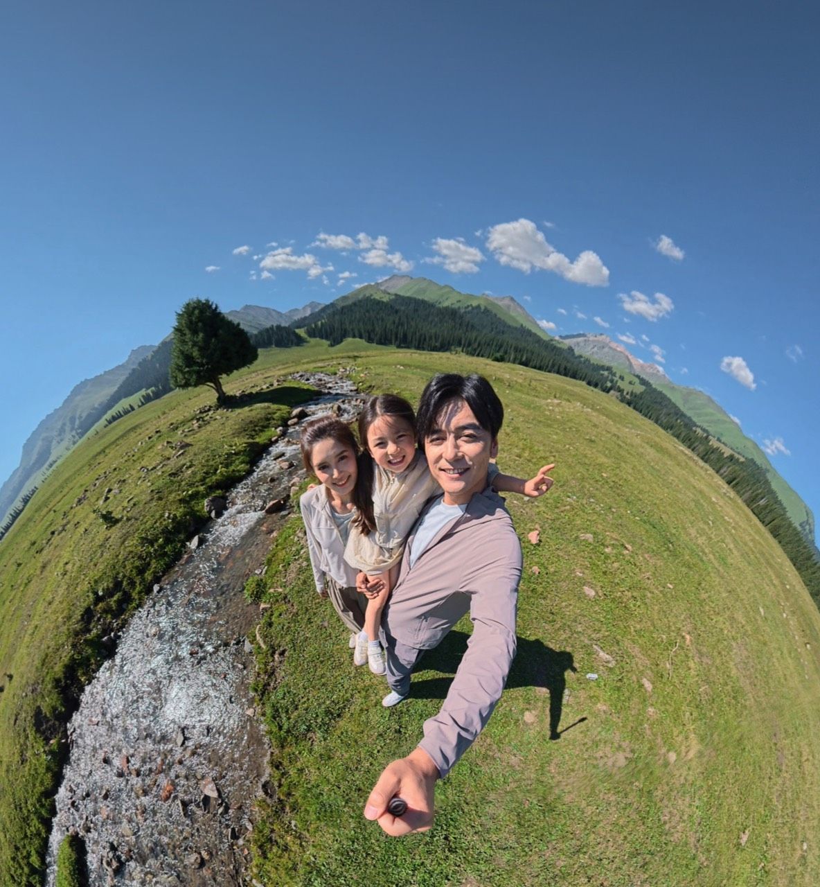 Three people smiling together in a scenic landscape with mountains and a stream.
