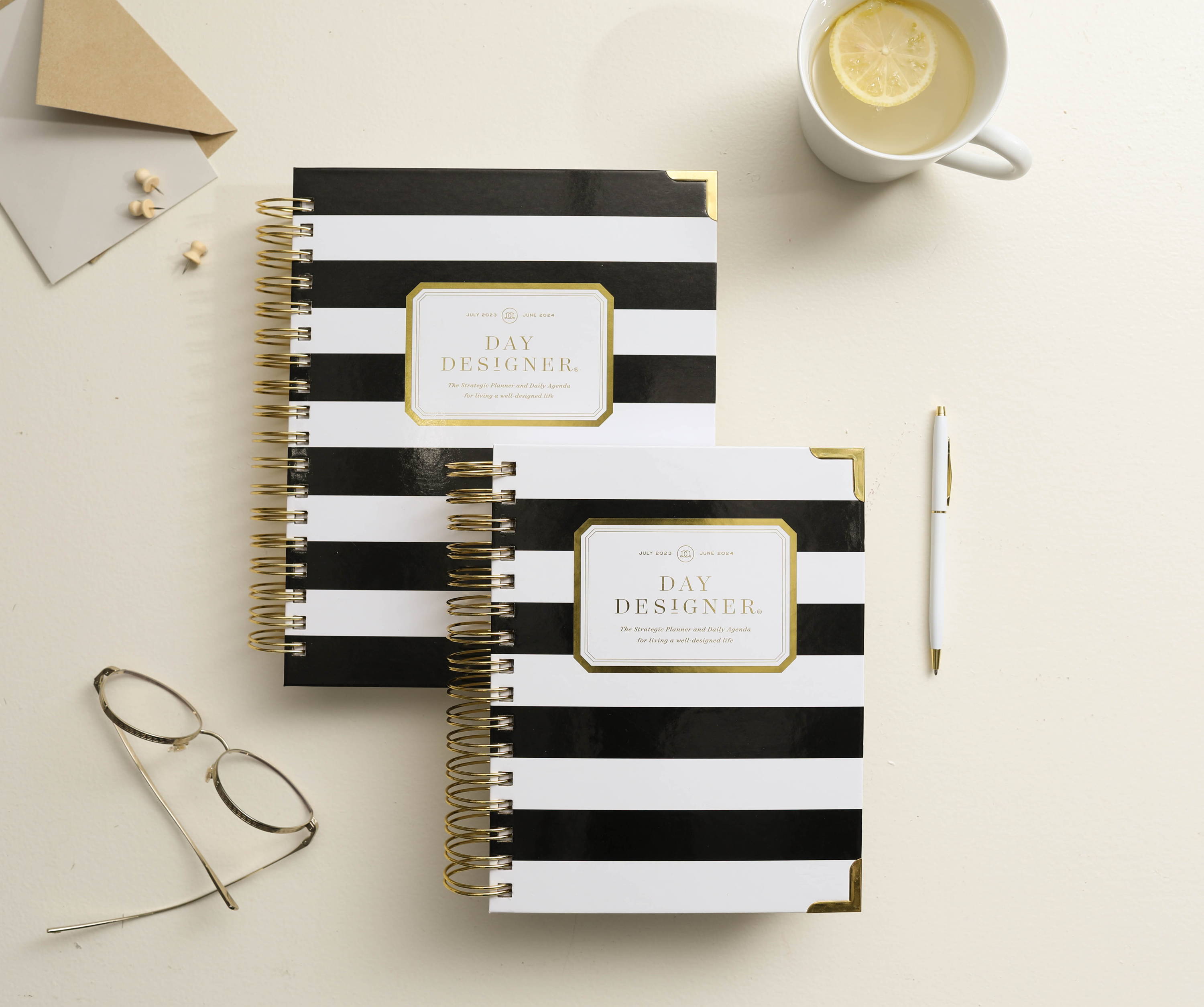 black and white planners, stacked to show sizing, white pen, reading glasses, mug with tea and lemon, notecard on beige background
