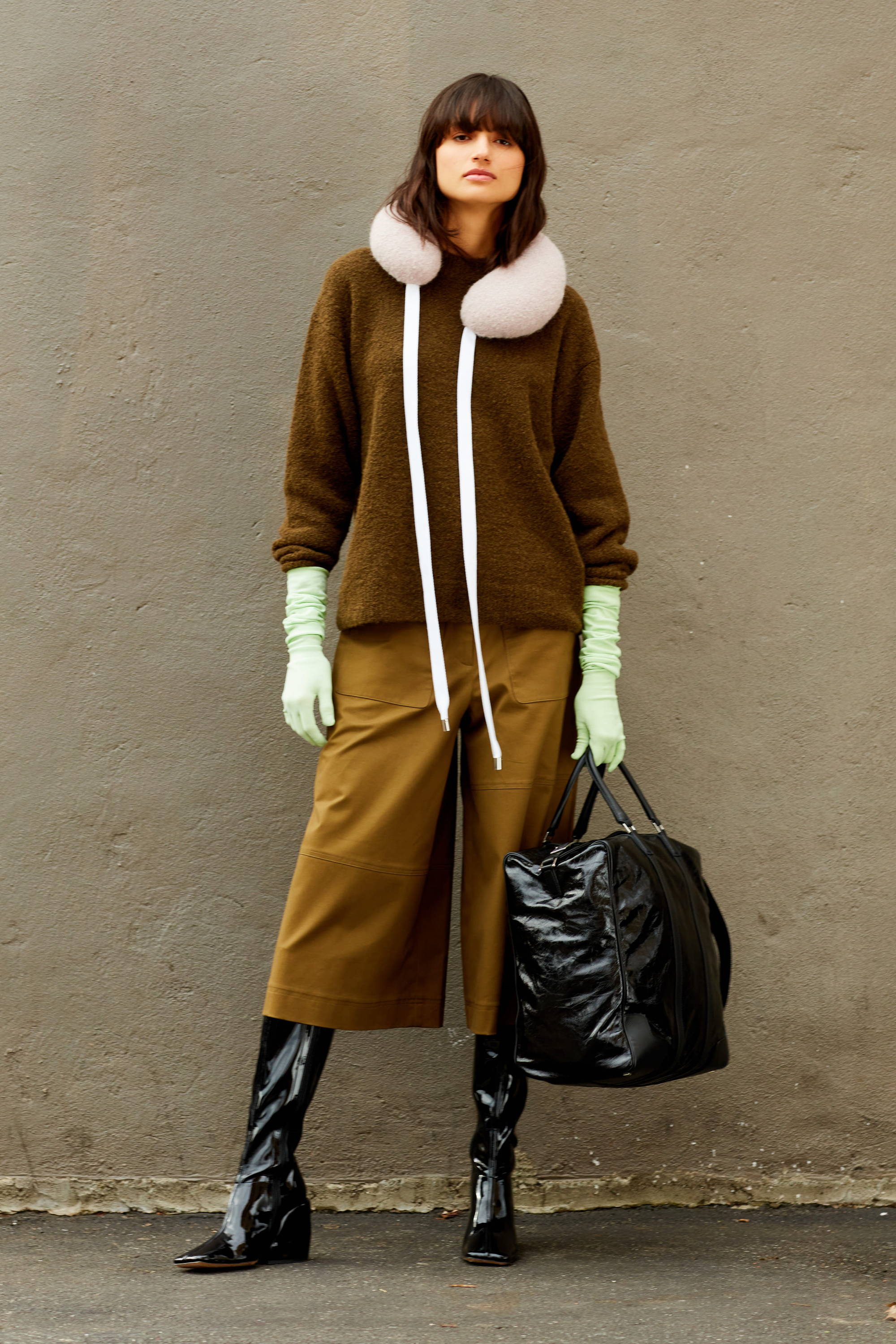 Woman in stylish outfit with gloves and a large bag against a neutral background.